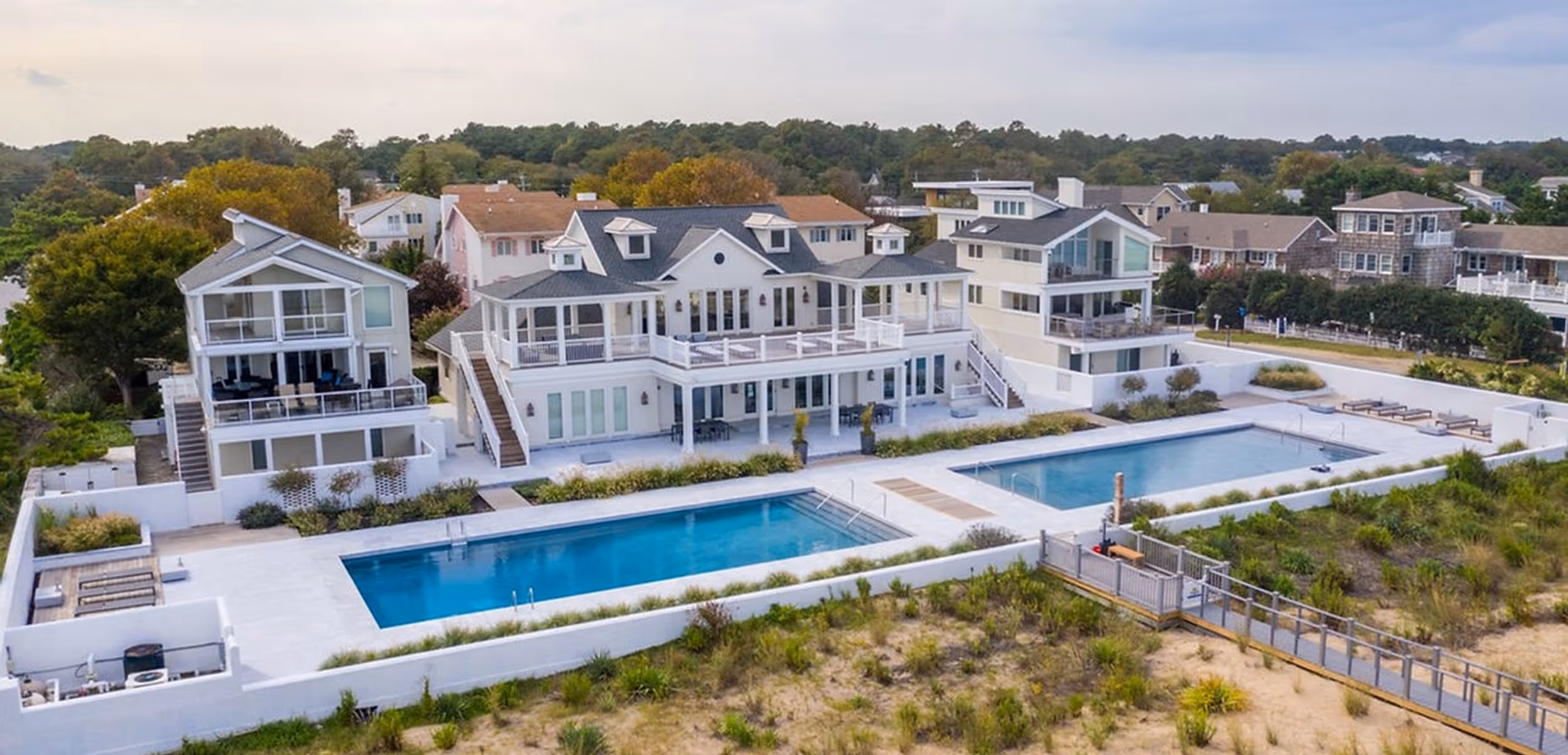 Large modern coastal mansion with two swimming pools surrounded by white walls and greenery.