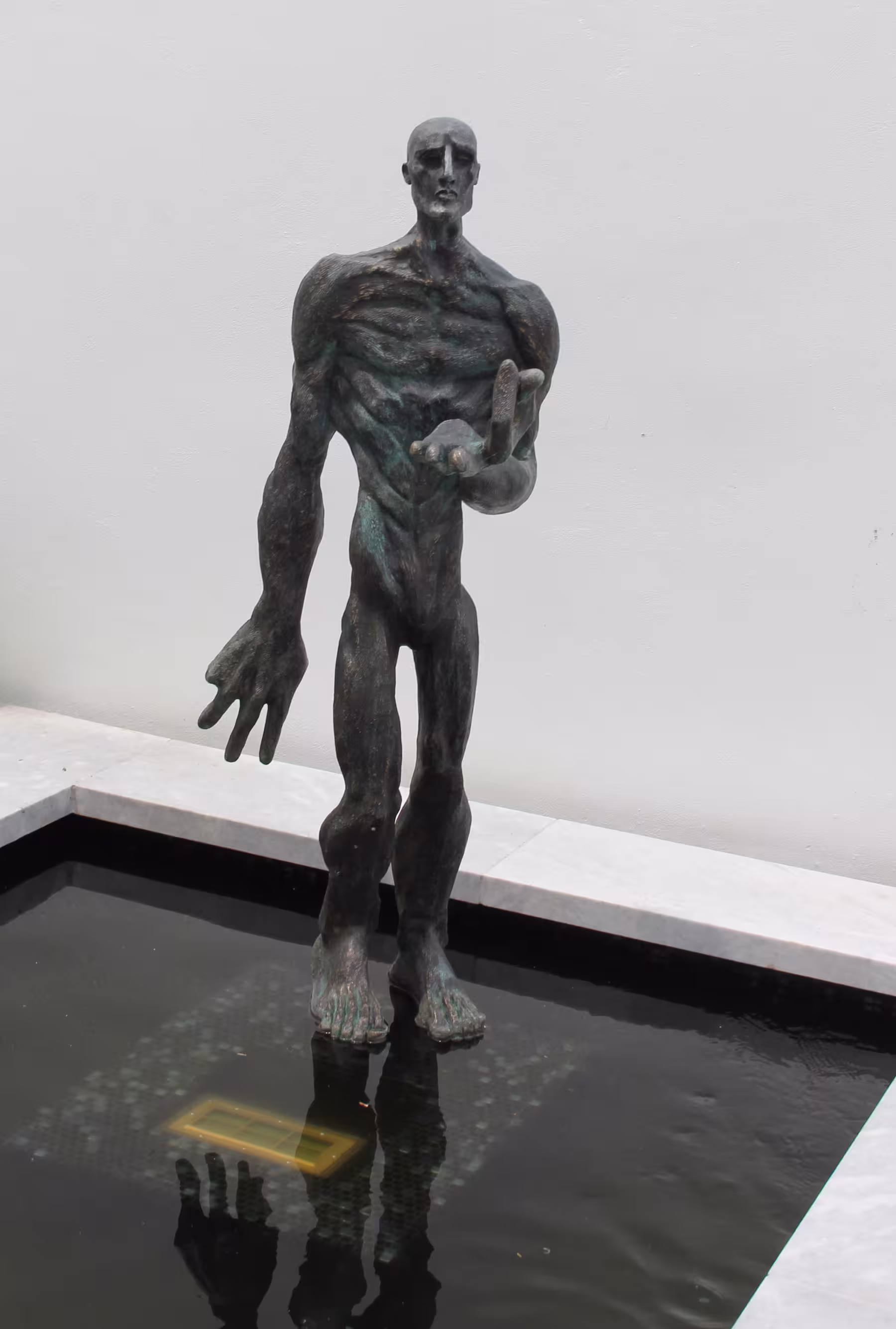 Abstract bronze sculpture of a muscular humanoid figure standing in a shallow black reflective pool against a plain white background.