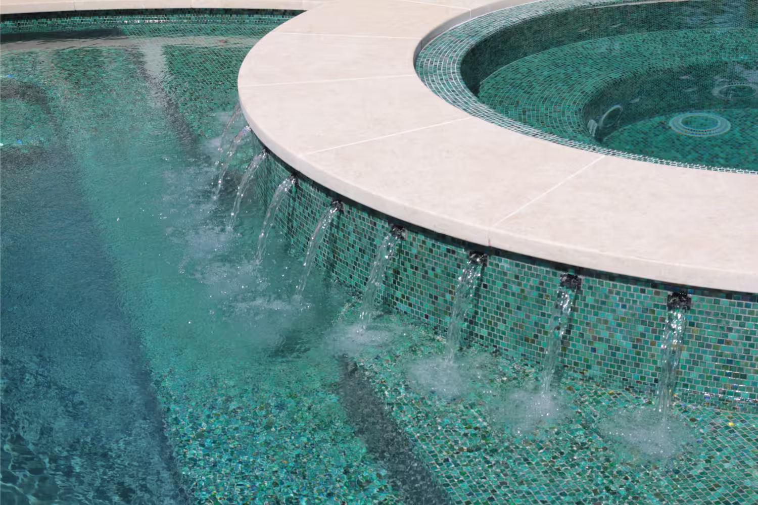 Circular spa with water jets flowing into a swimming pool lined with turquoise mosaic tiles.