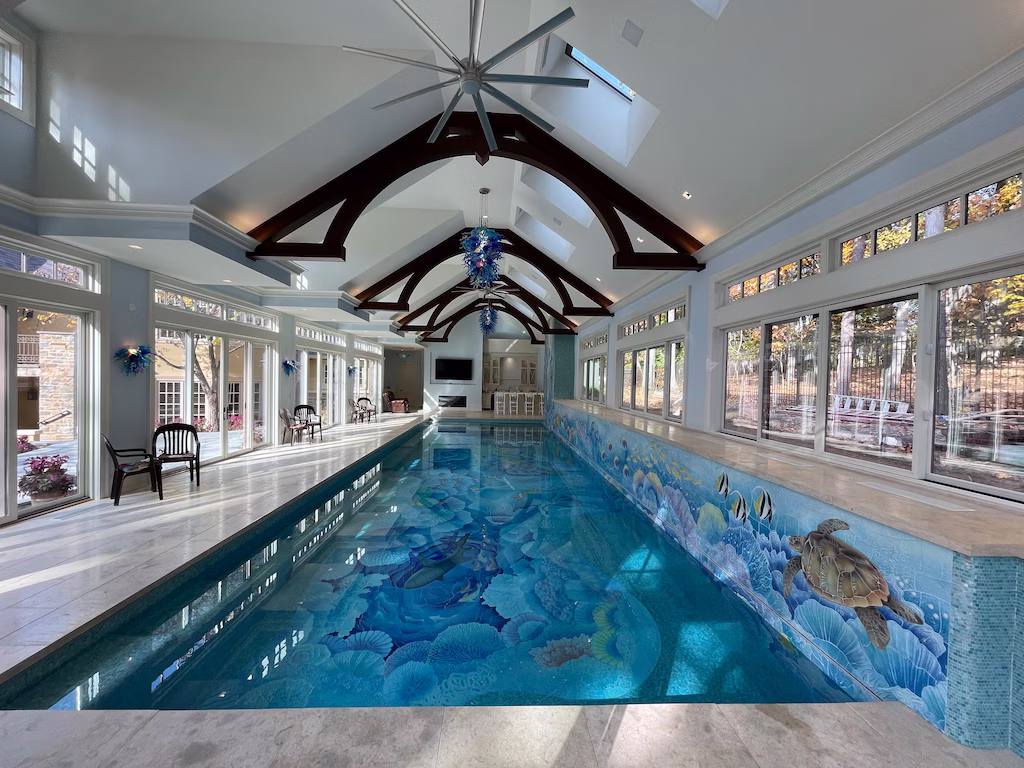 Indoor swimming pool with ocean-themed mosaic tiles featuring sea turtles and fish, large windows, and vaulted ceiling with dark wooden beams and ceiling fans.