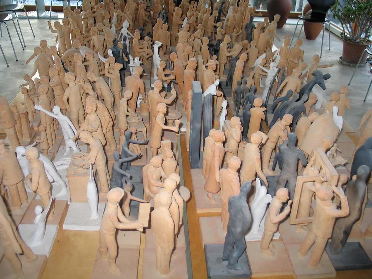 Viele kleine, farblich unterschiedliche Skulpturen von Menschen in verschiedenen Posen, auf Holzplatten arrangiert in einem hellen Raum mit Stühlen und Pflanzen im Hintergrund.