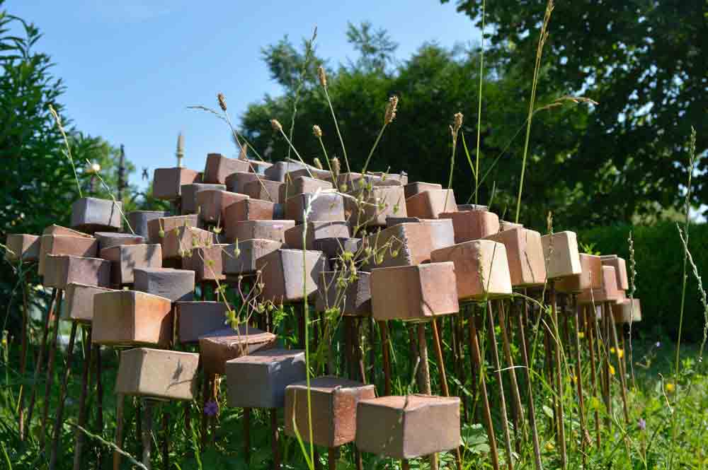 Kunstinstallation aus zahlreichen braunen und beigen Quadraten auf dünnen Metallstangen in einer grünen Wiese bei sonnigem Himmel.