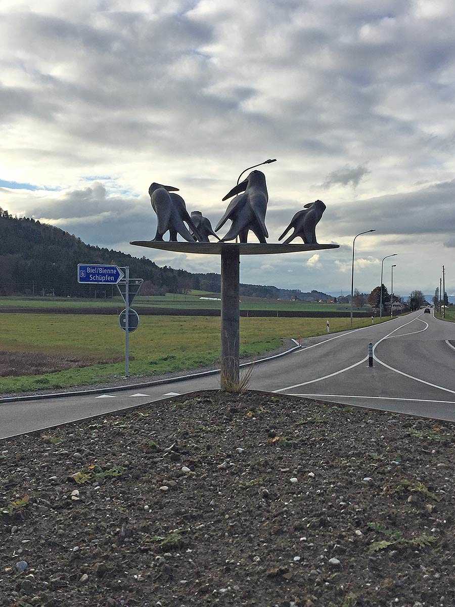 Rundverkehr mit einer Skulptur aus fünf grossen schwarzen Vögeln auf einer Plattform, ländliche Landschaft und Strassenschilder mit Richtung Biel/Bienne und Schüpfen im Hintergrund.