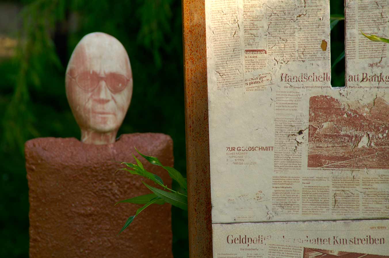 Skulptur eines menschlichen Kopfes mit Brille auf einem rustikalen Sockel neben einer verrosteten Metalltafel mit alten Zeitungsartikeln.