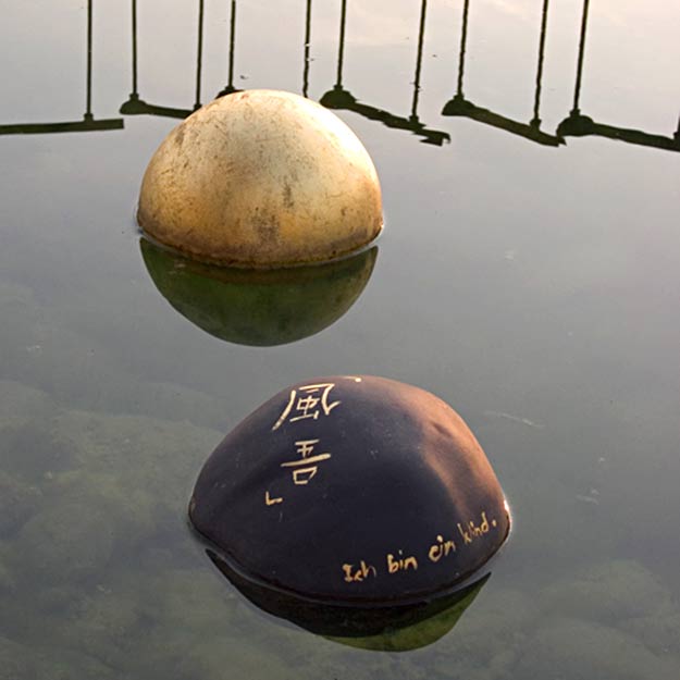 Zwei kugelförmige Objekte, eines hell und eines dunkel mit der Aufschrift ‚Ich bin ein Kind‘, schwimmen auf ruhigem Wasser mit Spiegelungen.
