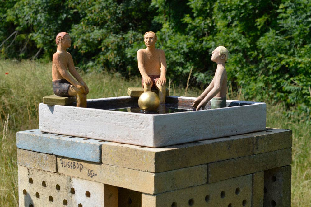 Drei kleine Figuren sitzen um ein quadratisches Wasserbecken mit einer goldenen Kugel darin, platziert auf einem gemauerten Sockel im Freien.