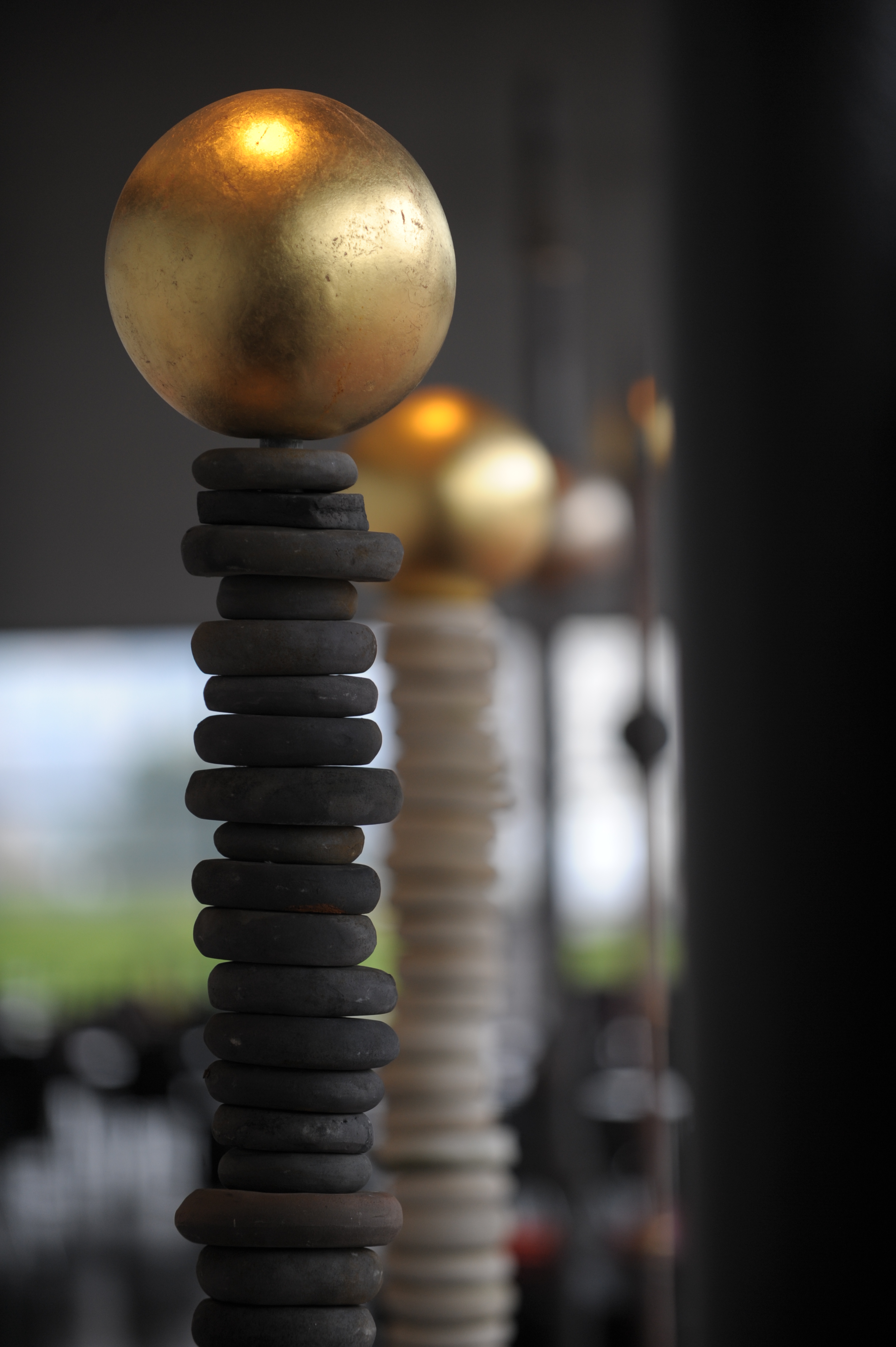 Skulptur aus aufgetürmten schwarzen Steinen mit einer goldenen Kugel darauf, unscharf im Hintergrund eine ähnliche Skulptur in Weiss.