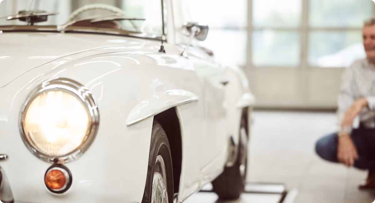 Weißer Oldtimer mit eingeschaltetem Scheinwerfer steht in einer hellen Werkstatt, während ein Gutachter das Fahrzeug aufmerksam begutachtet