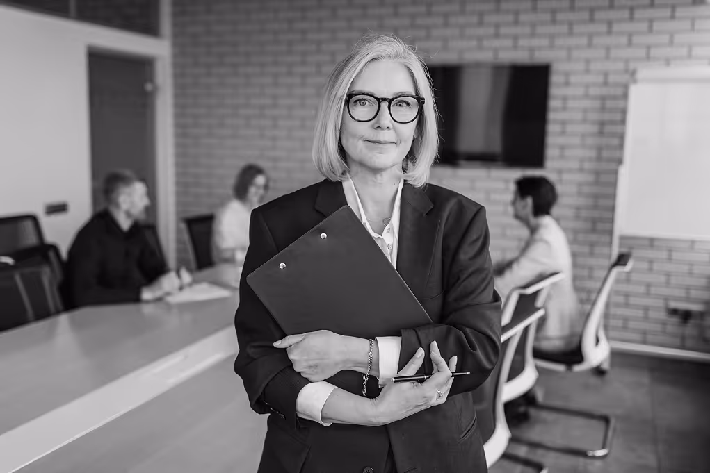 Woman in a conference room, smiling, with a meeting happening in the background