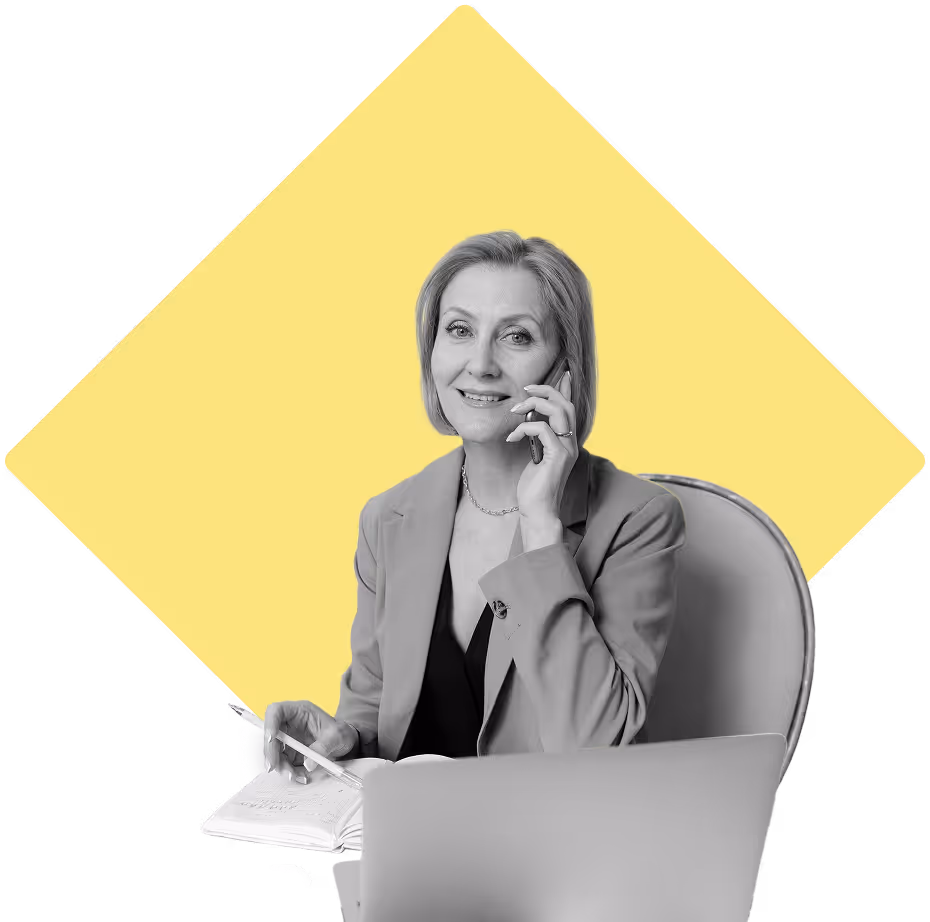 Woman sitting at her desk, smiling at the camera on the phone with a laptop, pen, and paper