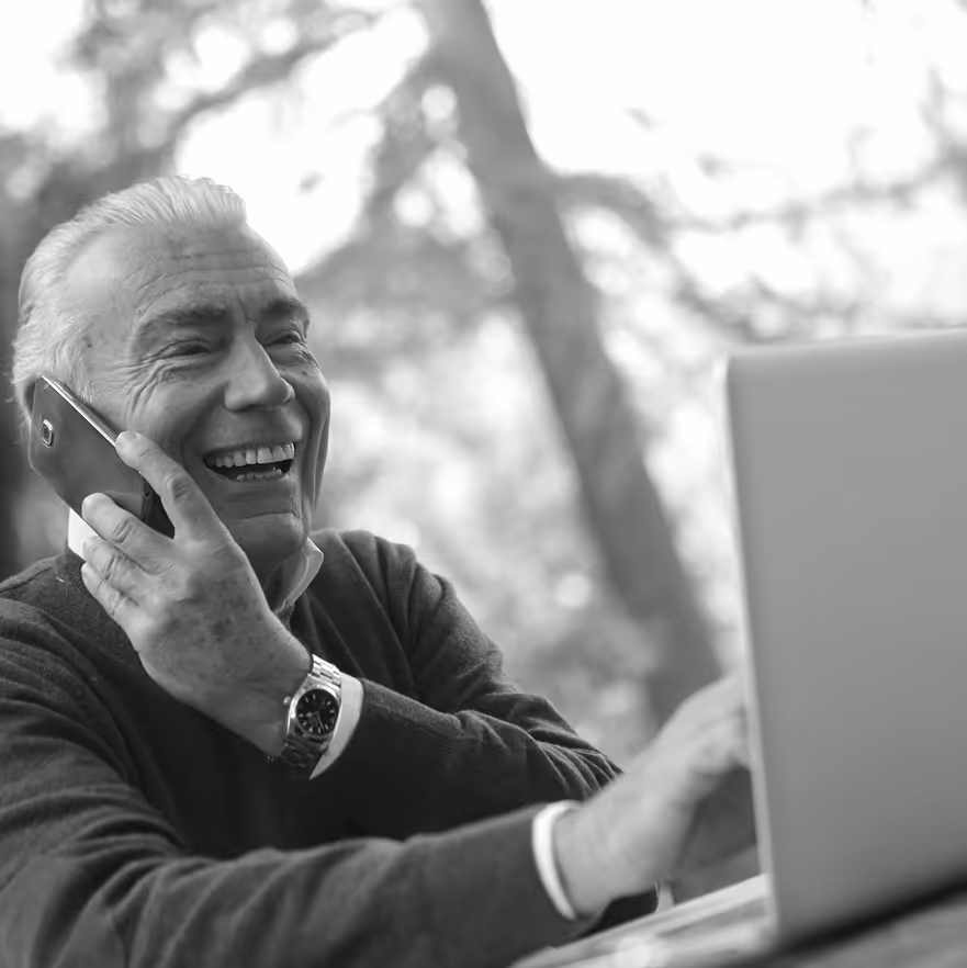 Older man speaking on his mobile phone while on his laptop outside.