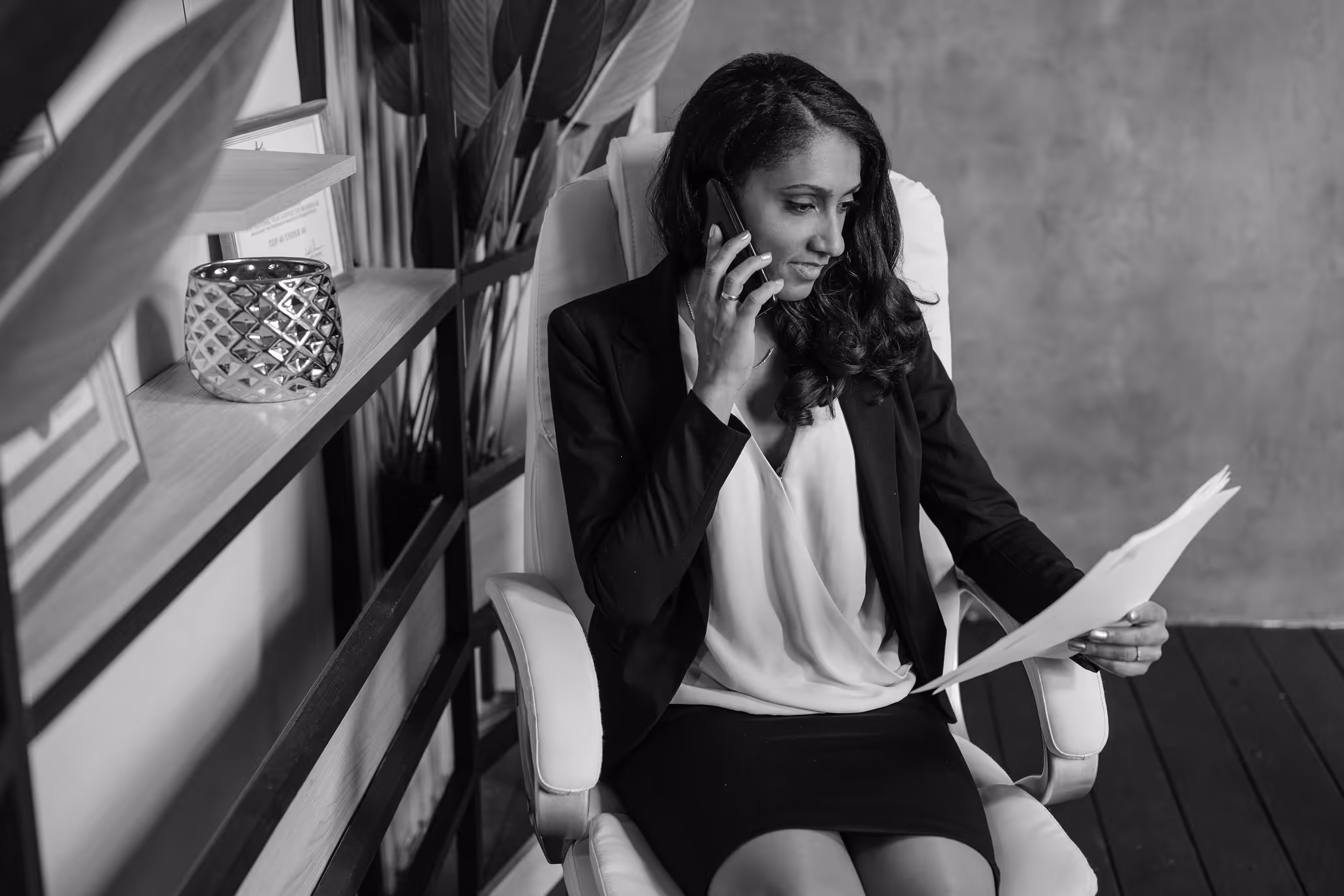 Woman in business attire sitting in an office chair, talking on phone and reviewing legal papers.