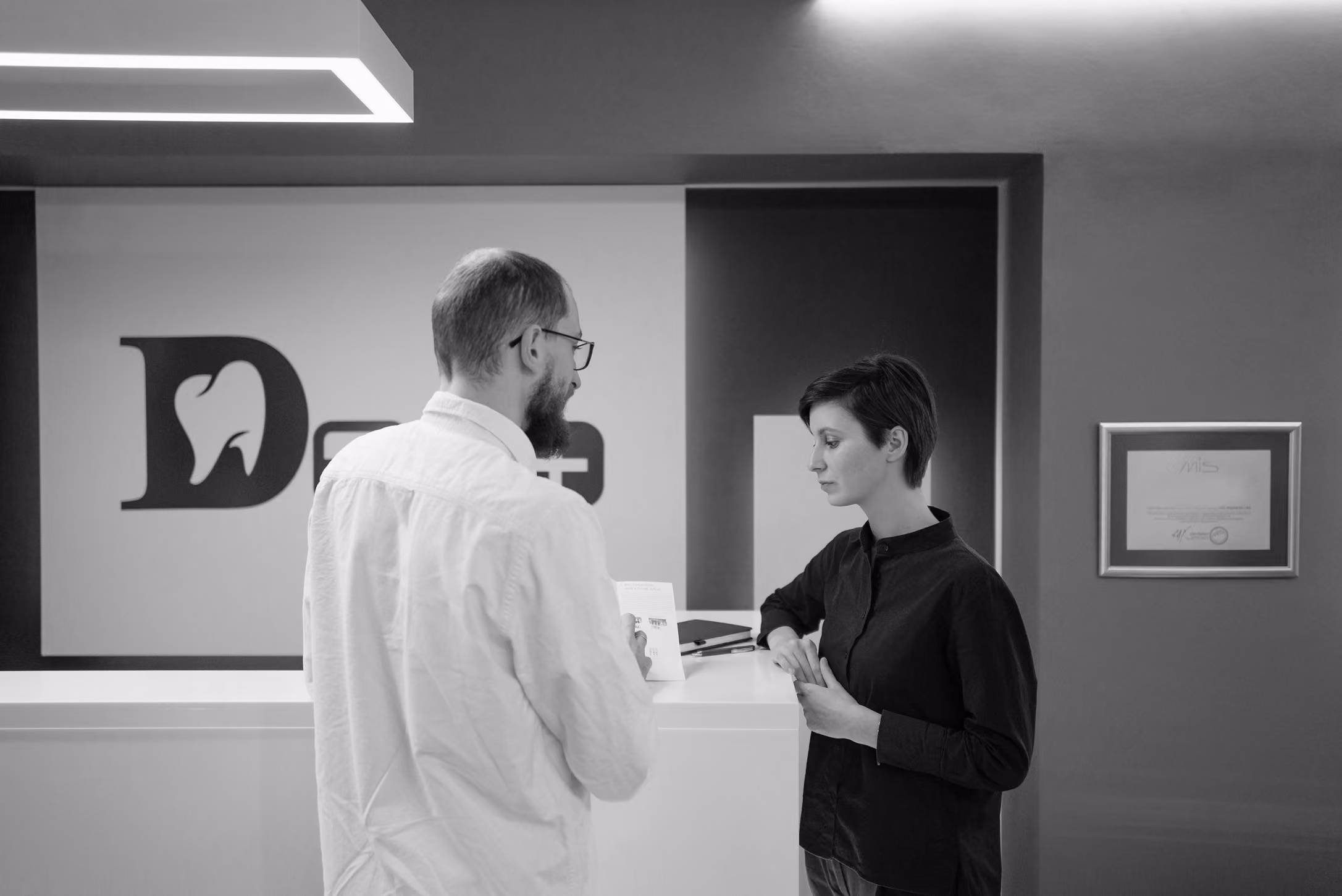 Dentist speaking to receptionist dental clinic's reception desk with tooth logo on the wall behind them.