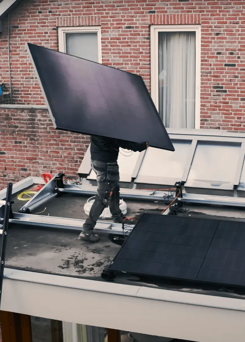 Een installateur tilt een groot zonnepaneel op een dak, waar meerdere zonnepanelen geïnstalleerd en gedeeltelijk zichtbaar zijn, tegen de achtergrond van een rode bakstenen huisgevel met ramen.