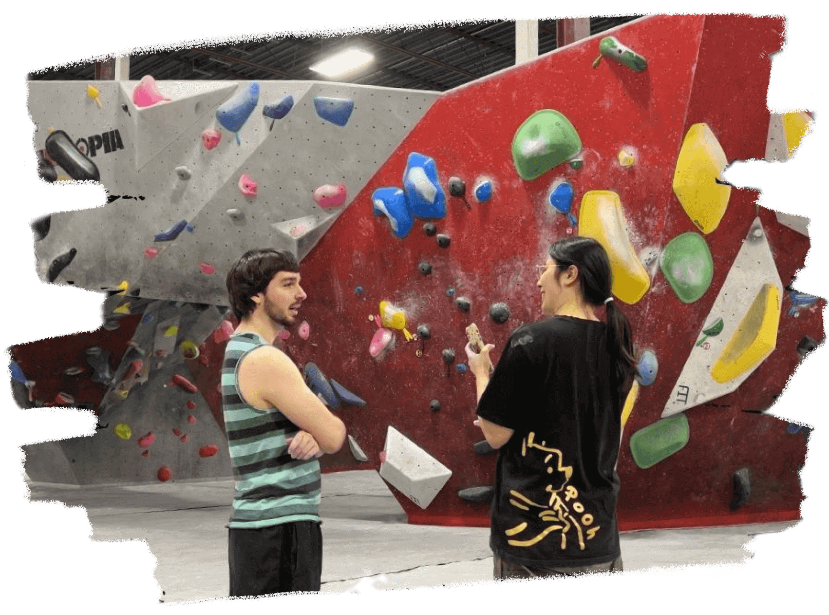 Product designer Lucy Lu conducting user research, interviewing a male climber in a bouldering gym. They are standing in front of a red and grey climbing wall with various colored holds.
