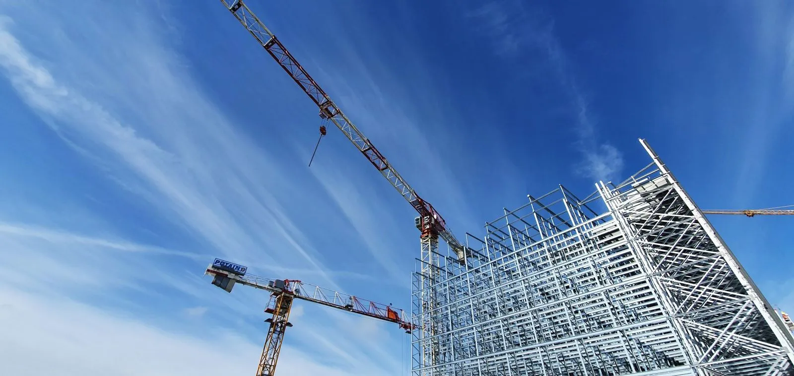 Construction site with cranes lifting materials and workers on elevated platforms near a large building structure.