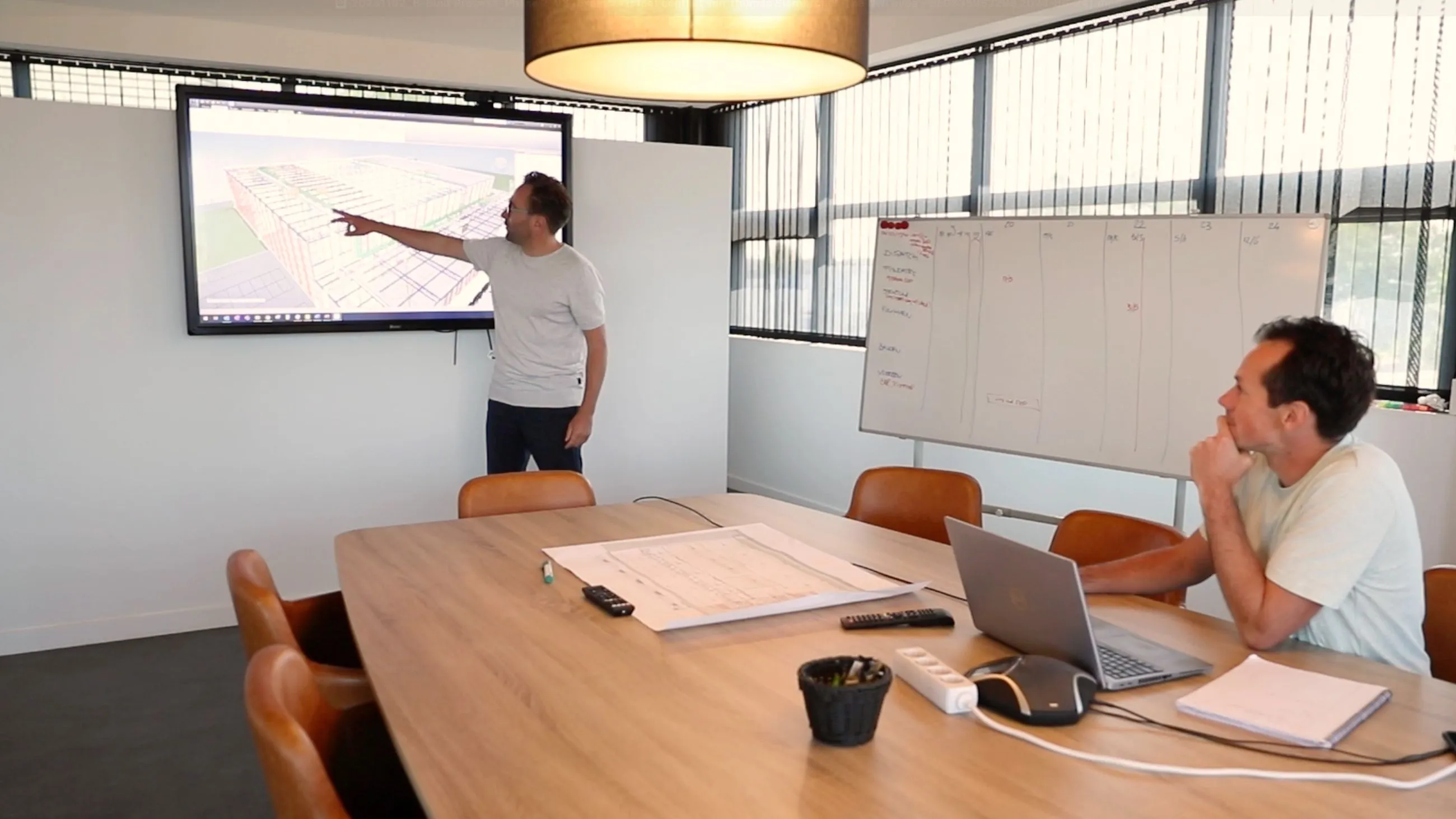 Two men in a meeting room discuss a map displayed on a large screen.