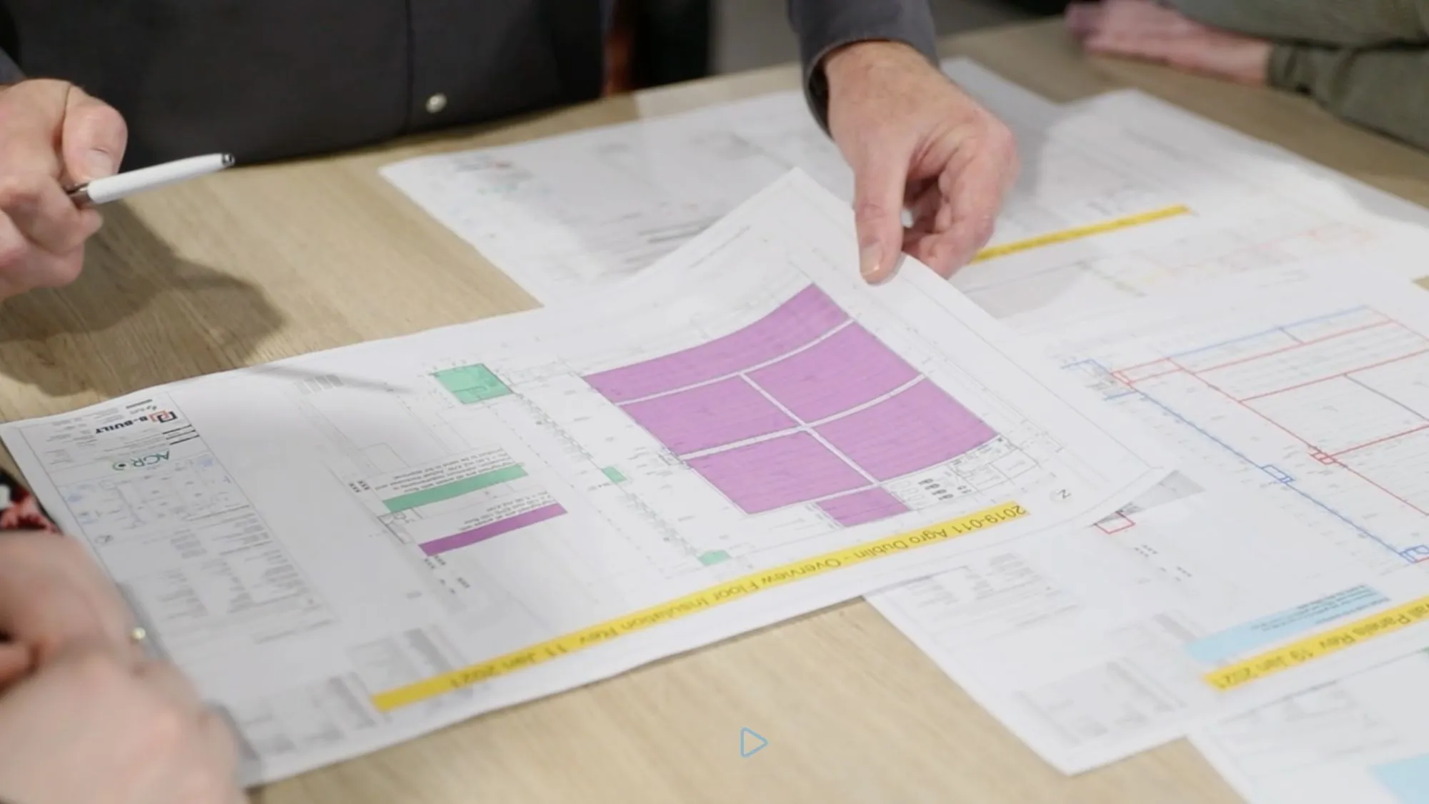 Hands hold and discuss architectural plans with pink highlighted areas on a wooden table.