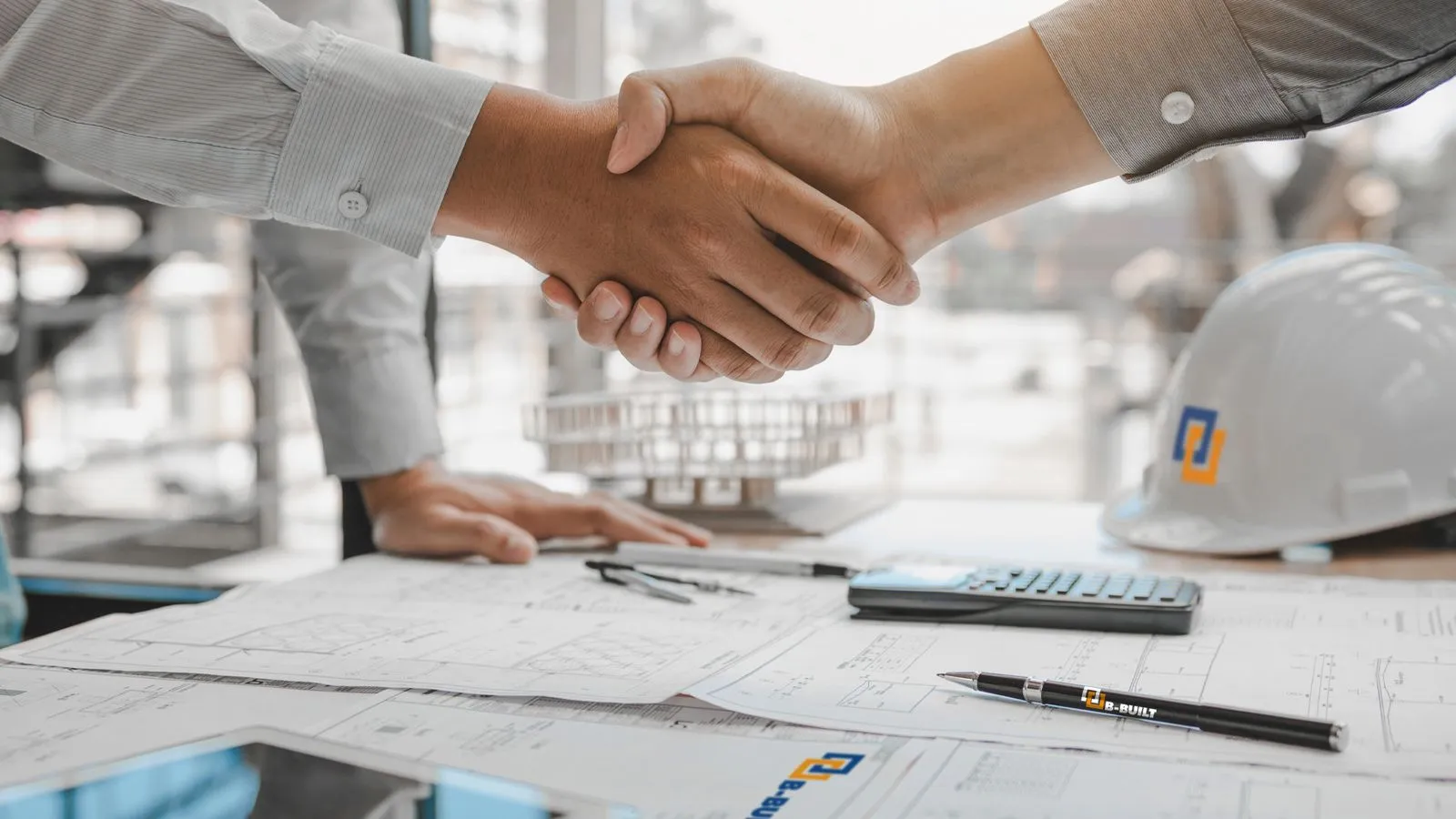 Two people shaking hands over architectural blueprints on a table, with a hard hat nearby.