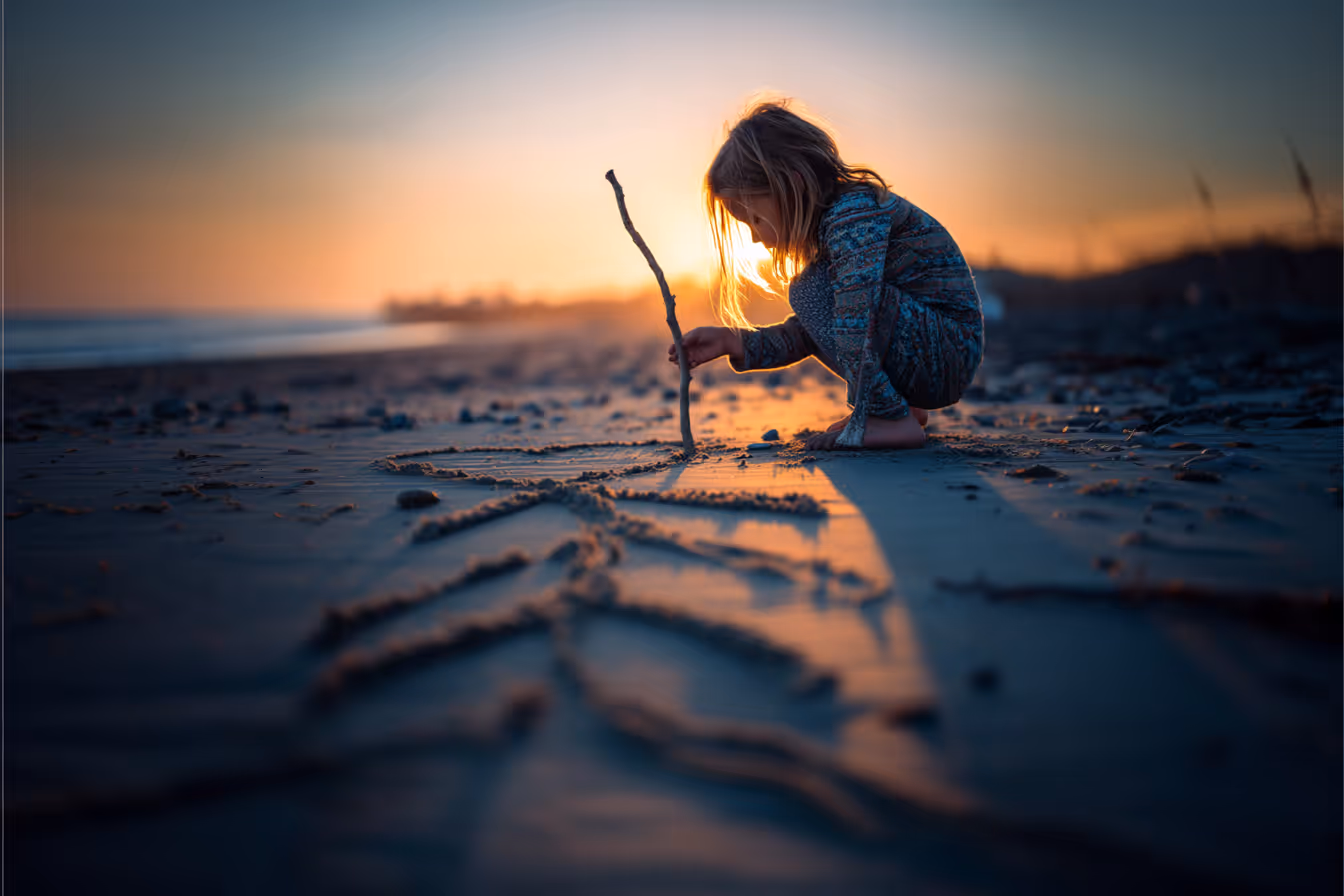 Child kneeling on the beach at sunset, drawing lines in the sand with a stick – a metaphor for intuitive creativity, simplicity, and pure expression.