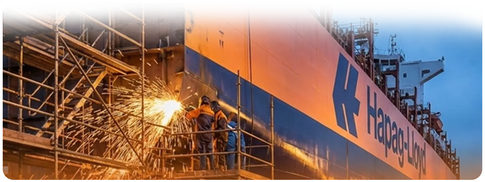 Close-up of Hapag-Lloyd vessel in a shipyard as workers produce sparks while welding beside heavy scaffolding.