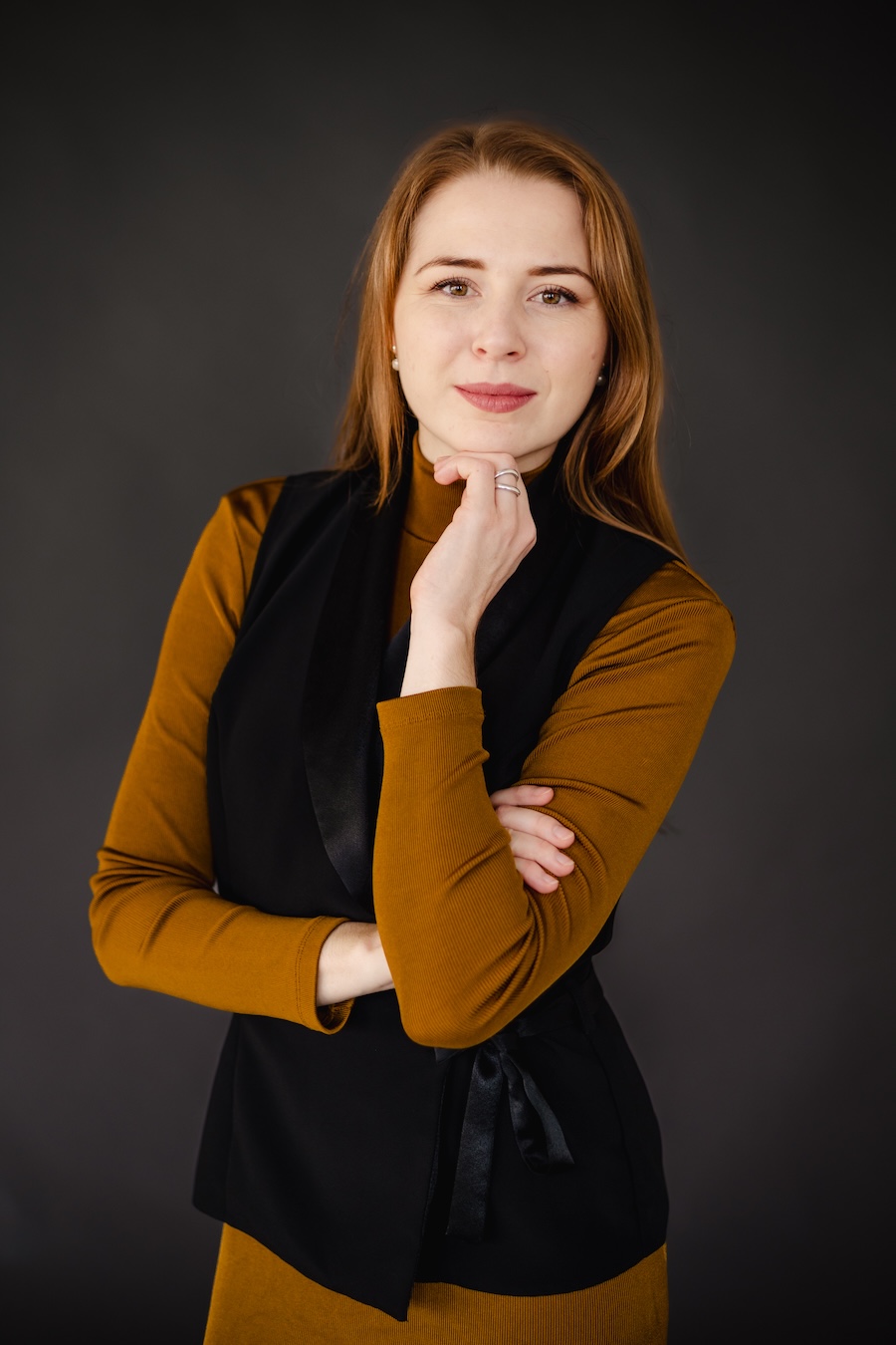Young woman with red hair wearing a mustard turtleneck and black vest posing thoughtfully against a dark background.