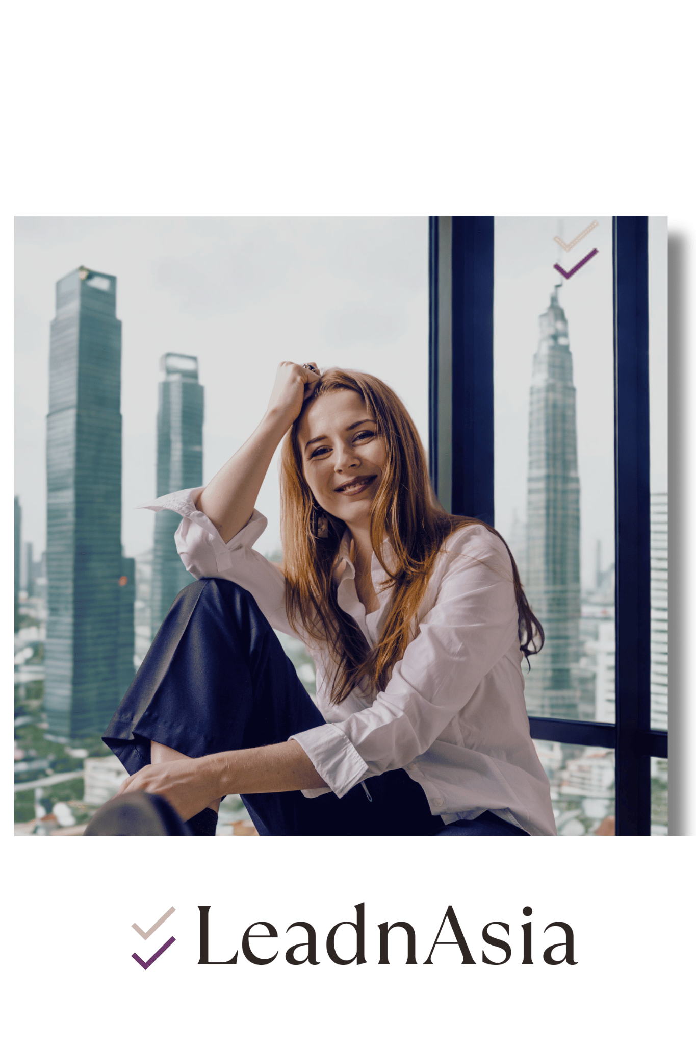 Smiling woman with red hair sitting by a large window overlooking a city skyline with tall skyscrapers.