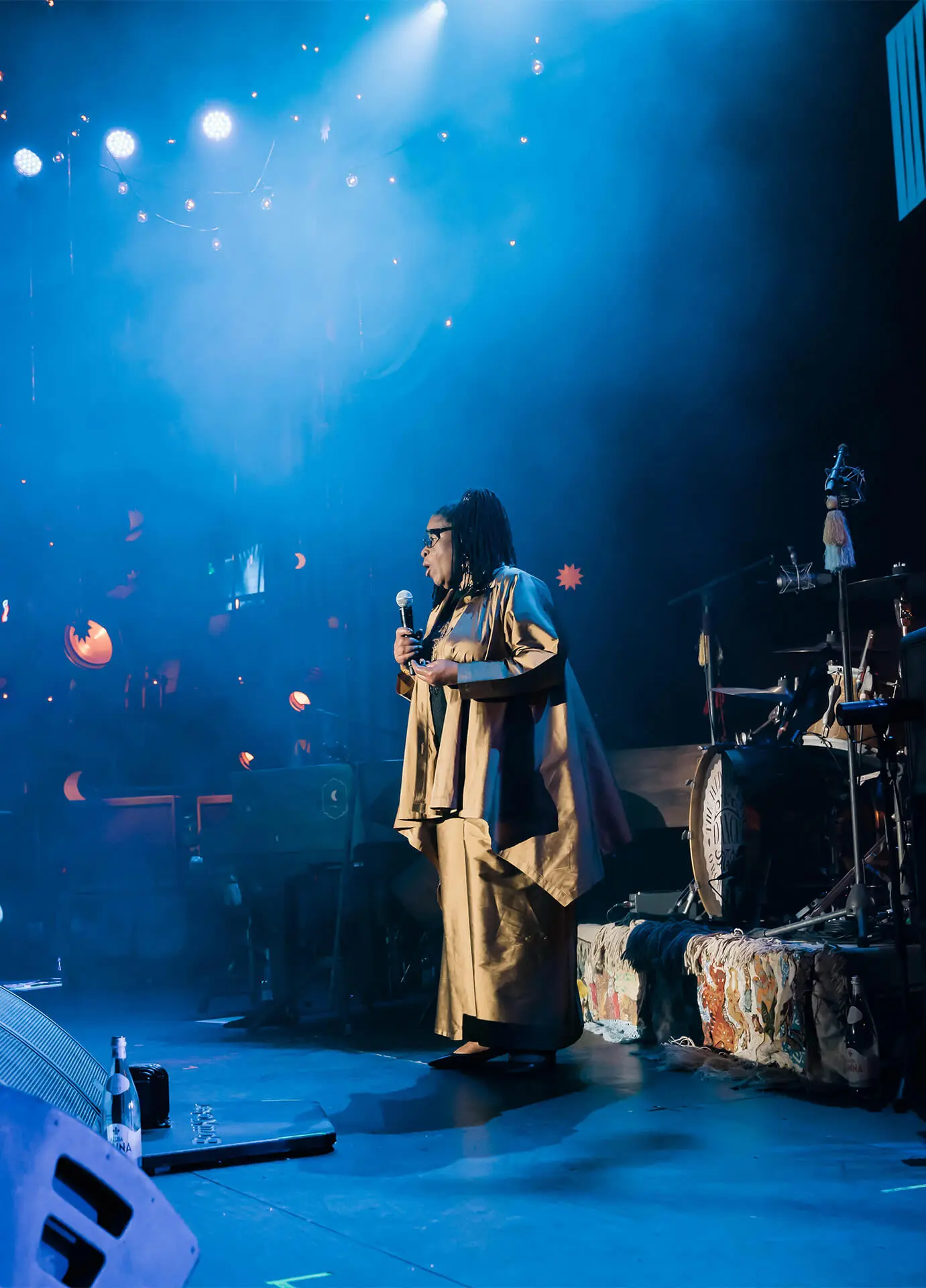 Woman in tan outfit speaking with microphone on stage with dramatic blue lighting at A New Way of Life Reentry Project event, a Los Angeles-based organization dedicated to supporting formerly incarcerated women