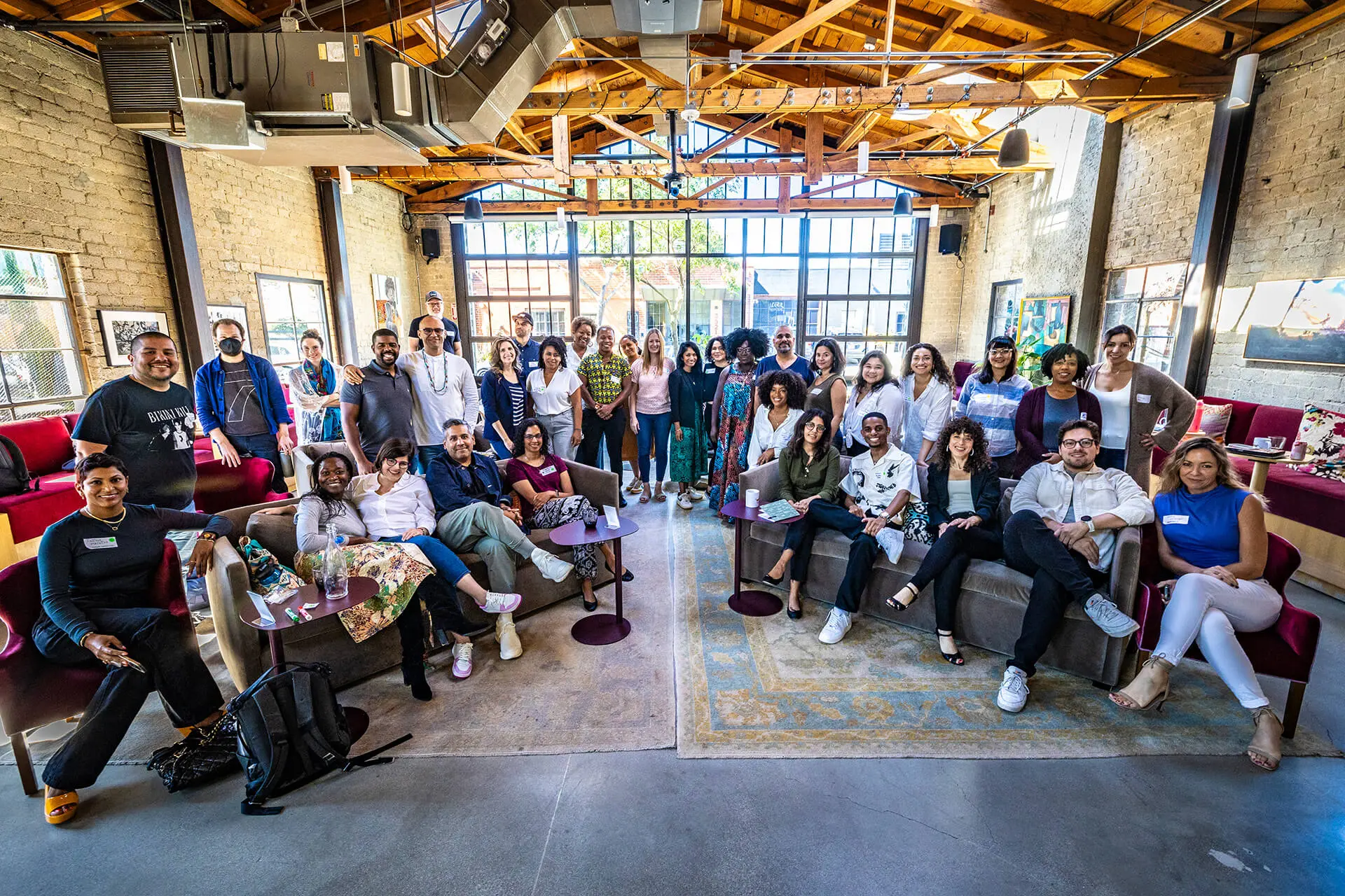 Large group photo of diverse attendees gathered in industrial-style event space with exposed brick walls, wooden beams, and modern seating areas