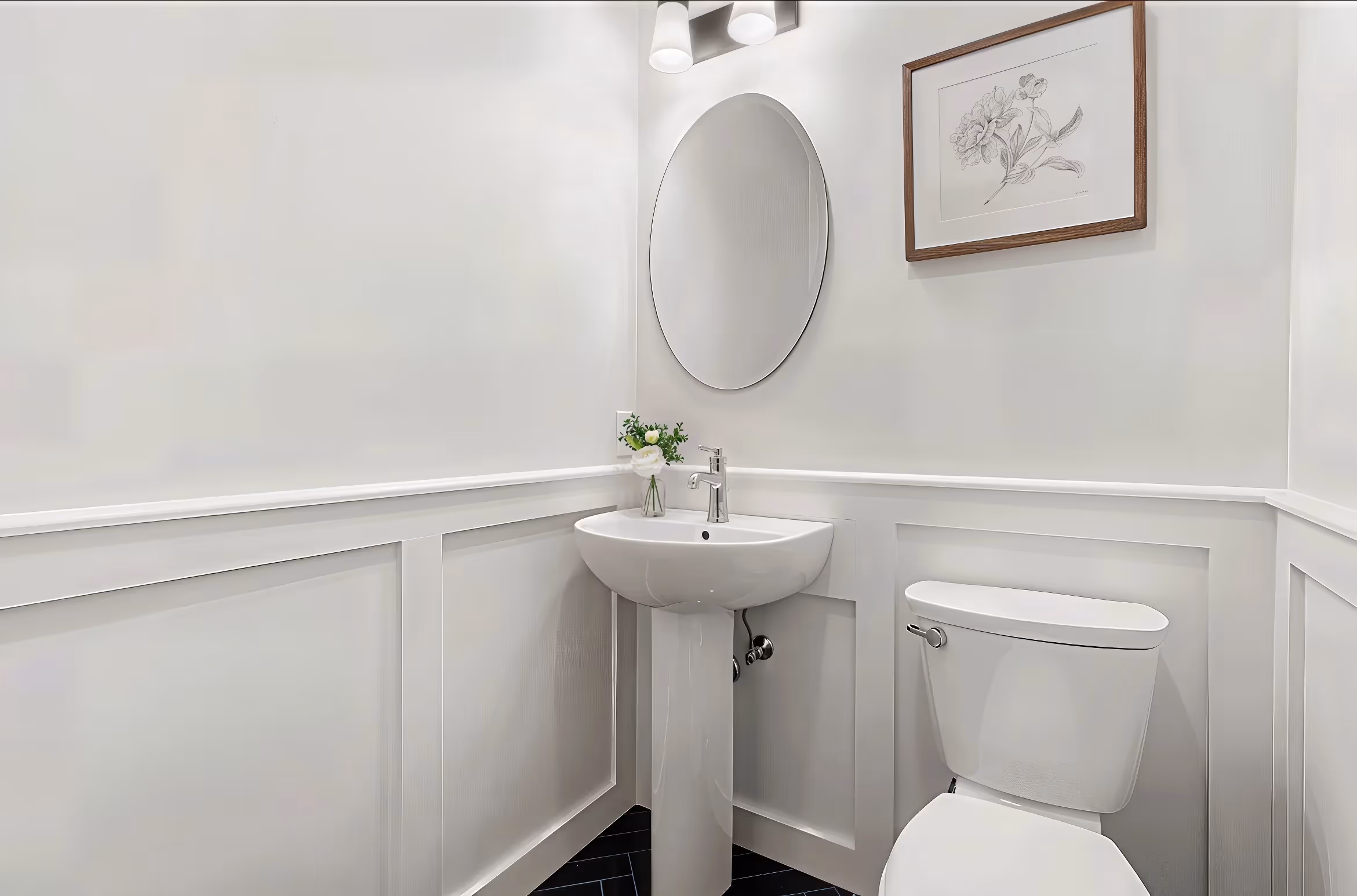 Small white bathroom with pedestal sink, round mirror, toilet, and framed flower art on the wall.