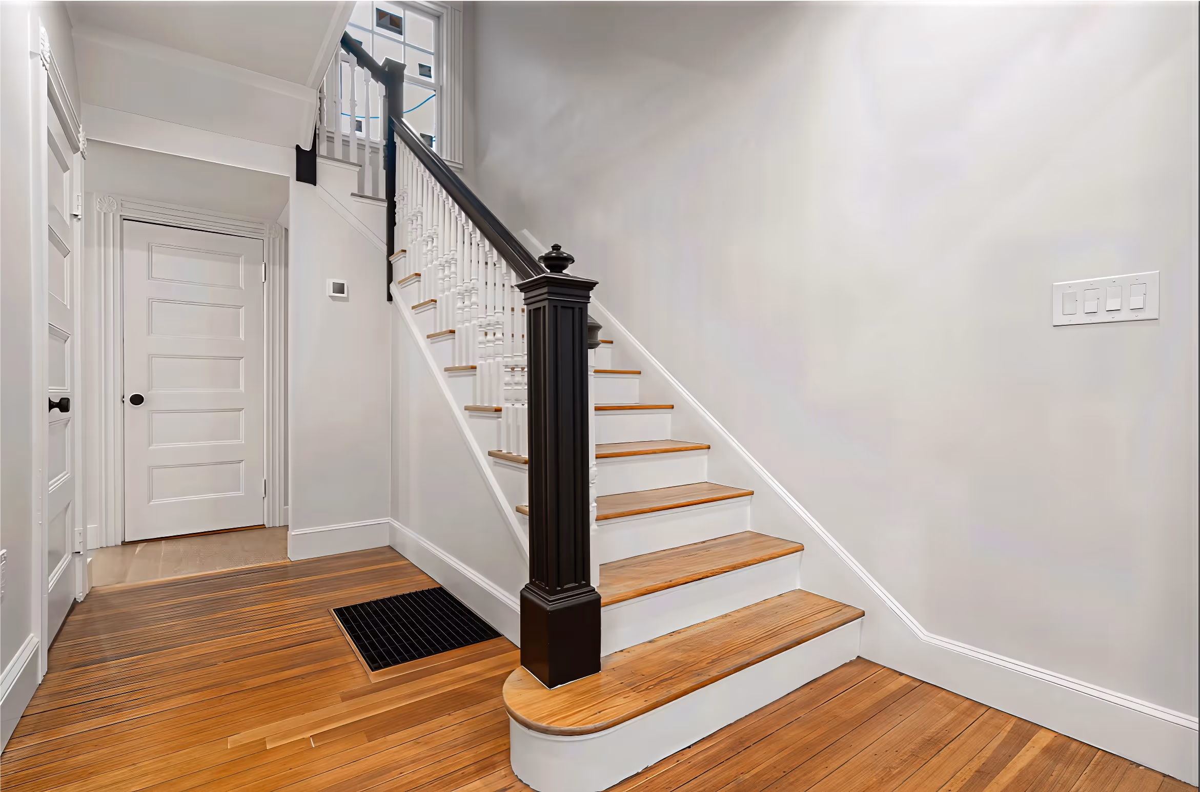 A wooden staircase with white risers and a dark railing in a hallway with hardwood floors and white walls.