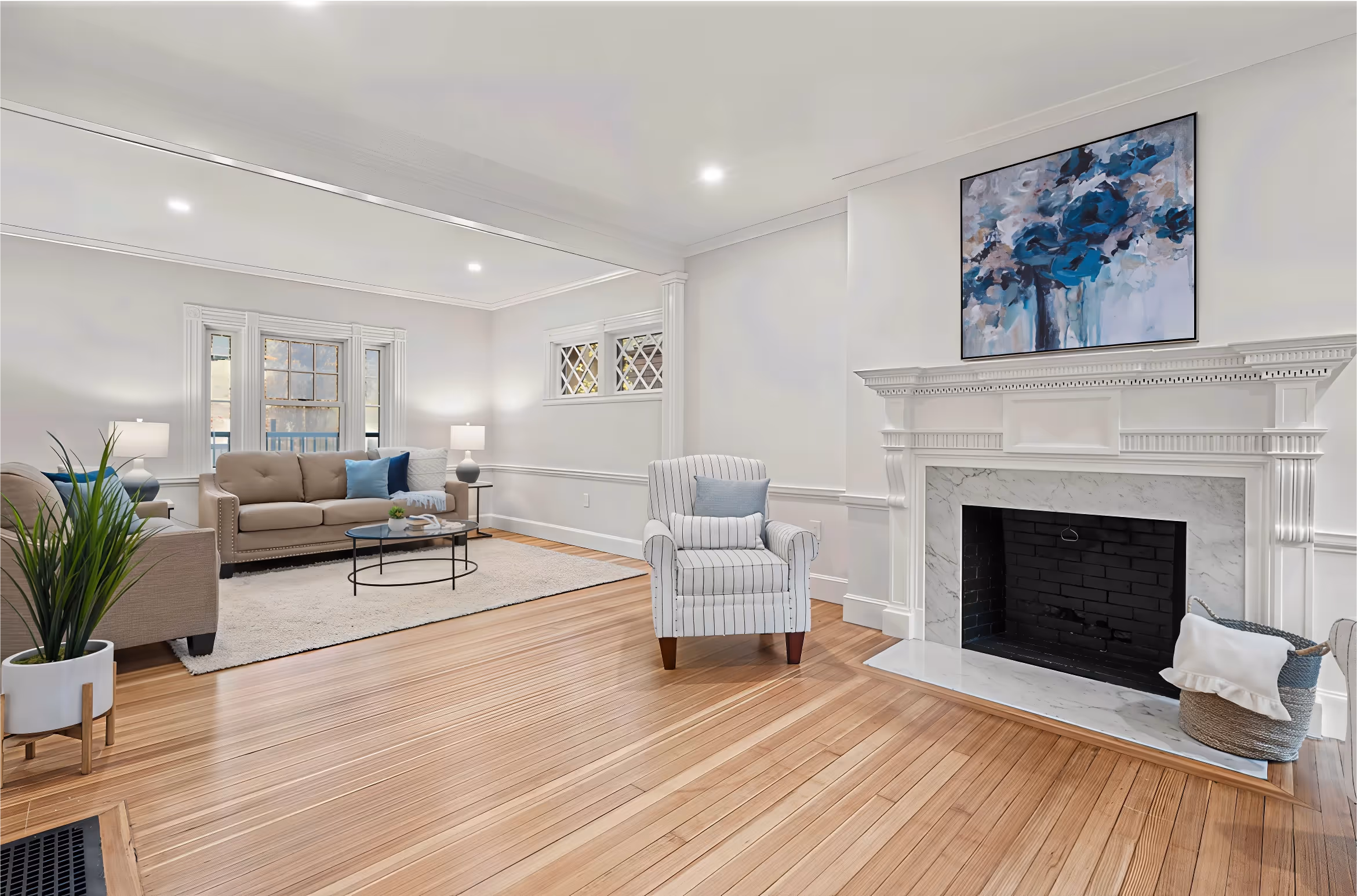 Bright living room with hardwood floors, a marble fireplace, sofas, armchair, and large floral painting above mantel.