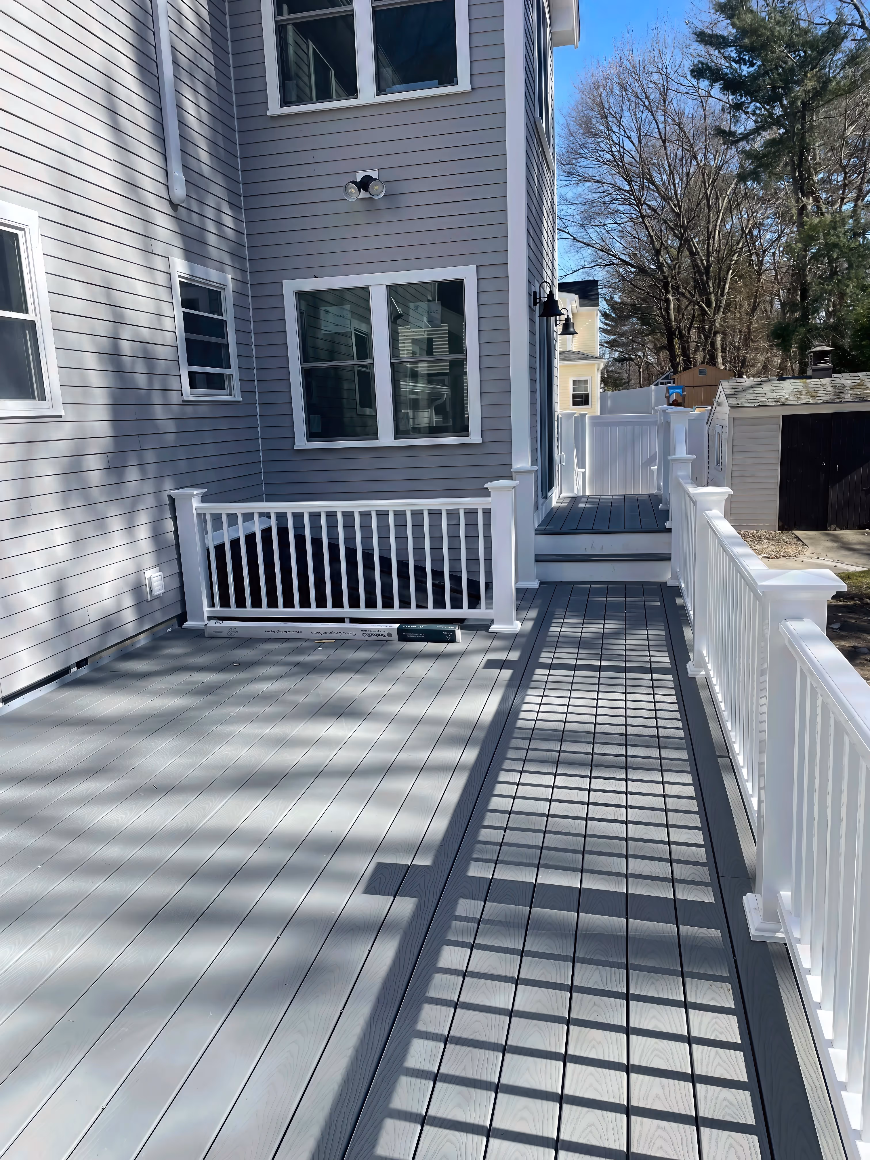 A gray deck with white railings sits next to the house, leading to a gate and backyard on a sunny day.