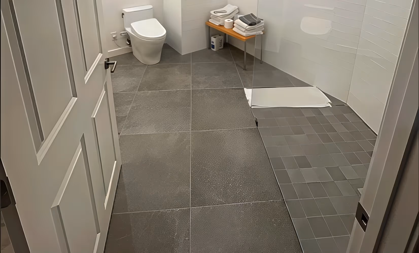 Modern bathroom with gray shower tile floor, toilet, walk-in shower, bench with towels, and a white bath mat.
