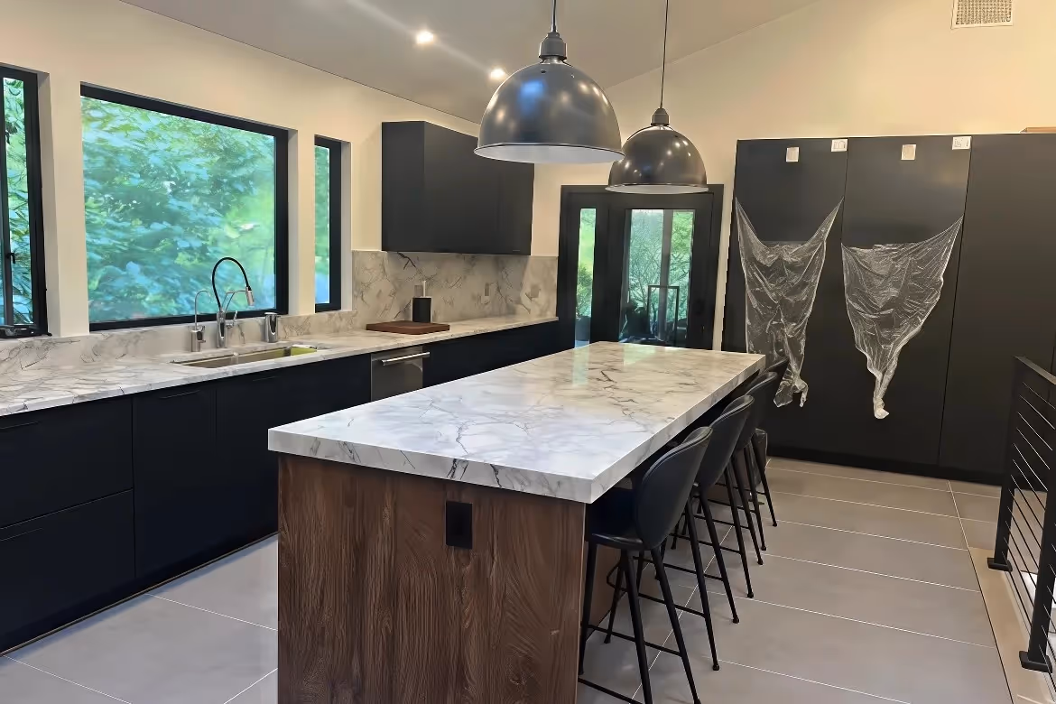 Modern kitchen with black cabinets, marble countertops, island seating, and large windows with a wooded view.