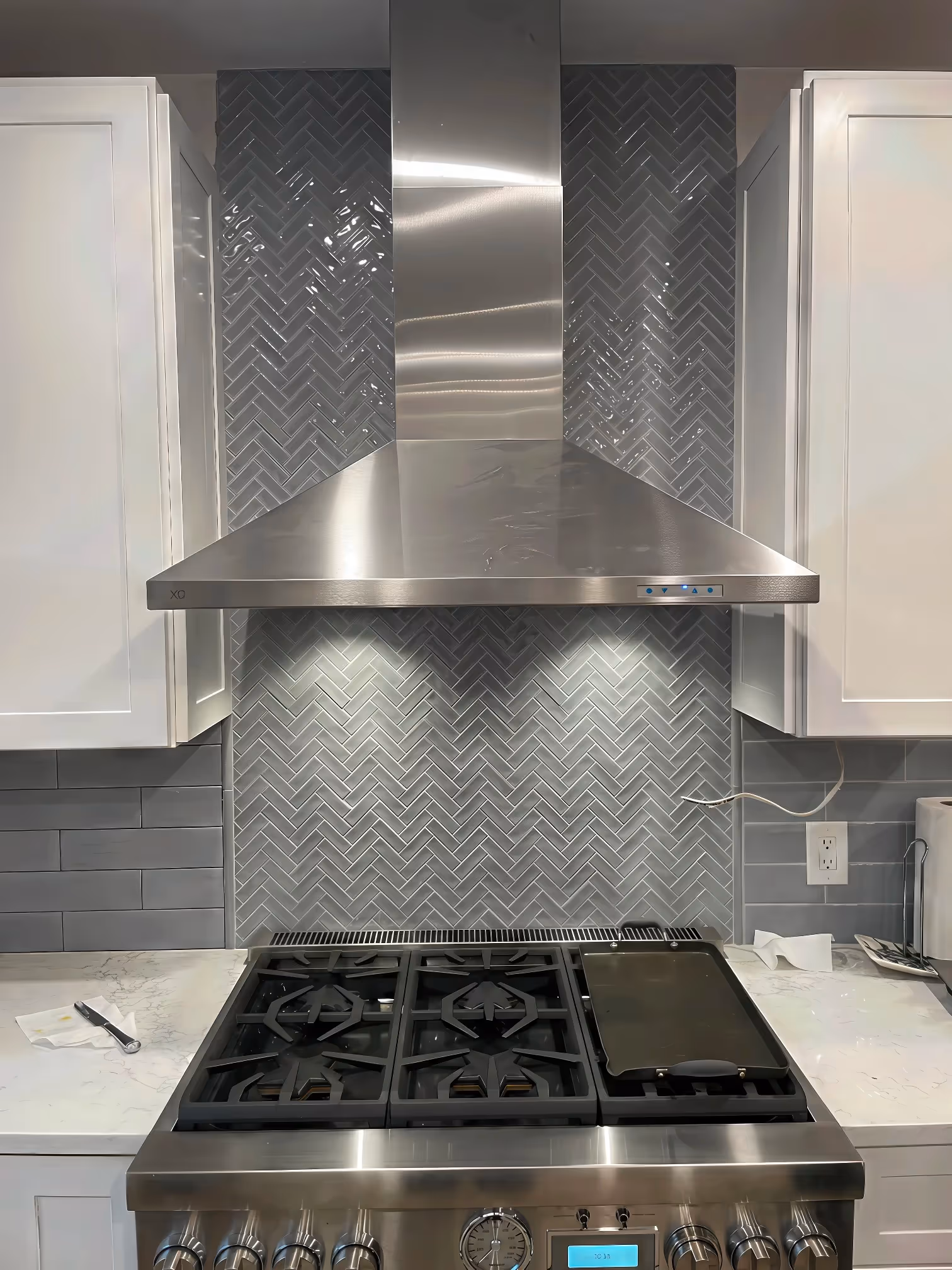 Stainless steel range hood over stove with gray herringbone tile backsplash, white cabinets, and high-end finishes.