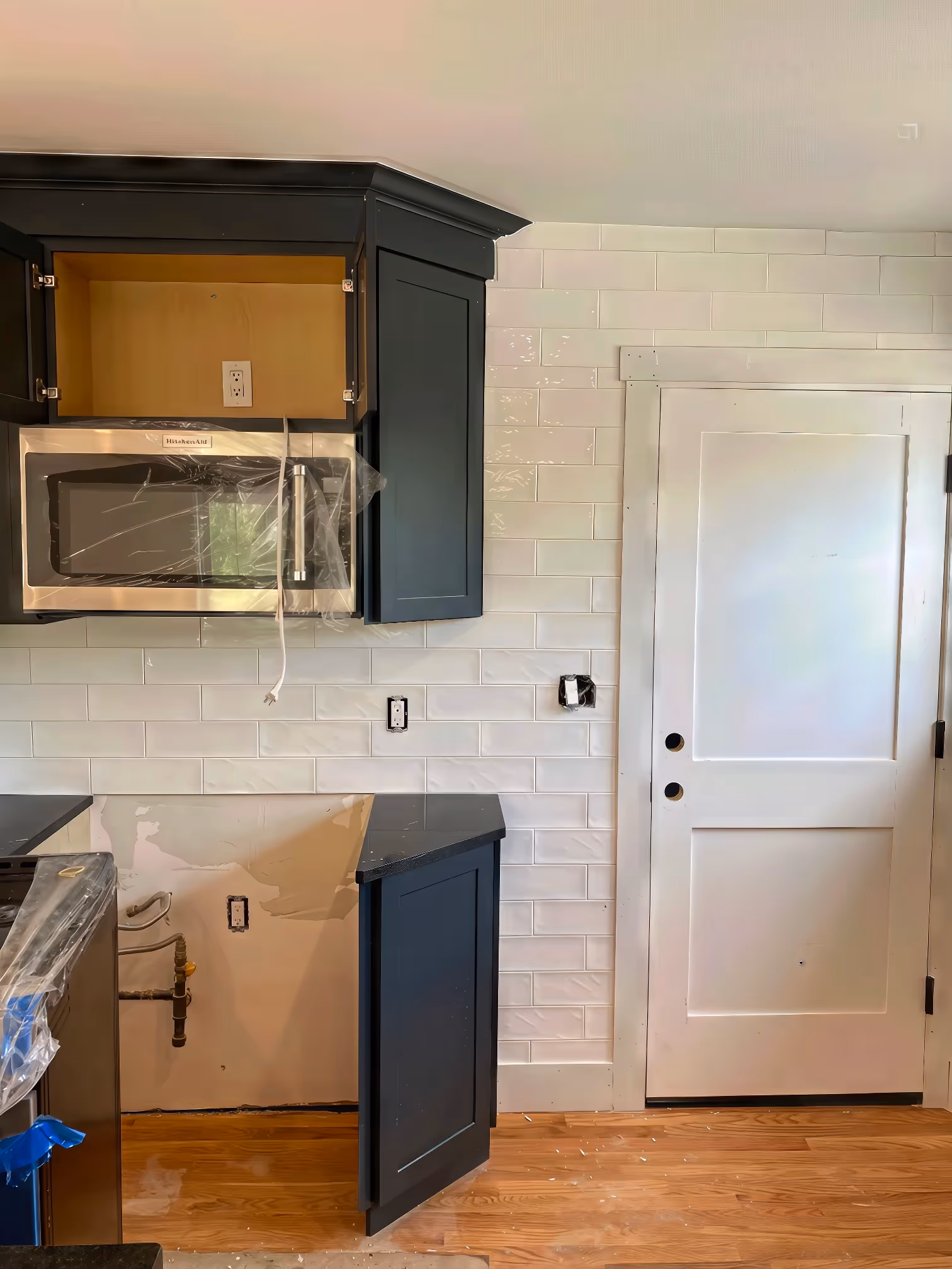Kitchen under renovation in Boston featuring custom cabinetry, dark cabinets, a microwave, white tile backsplash, and a white door.