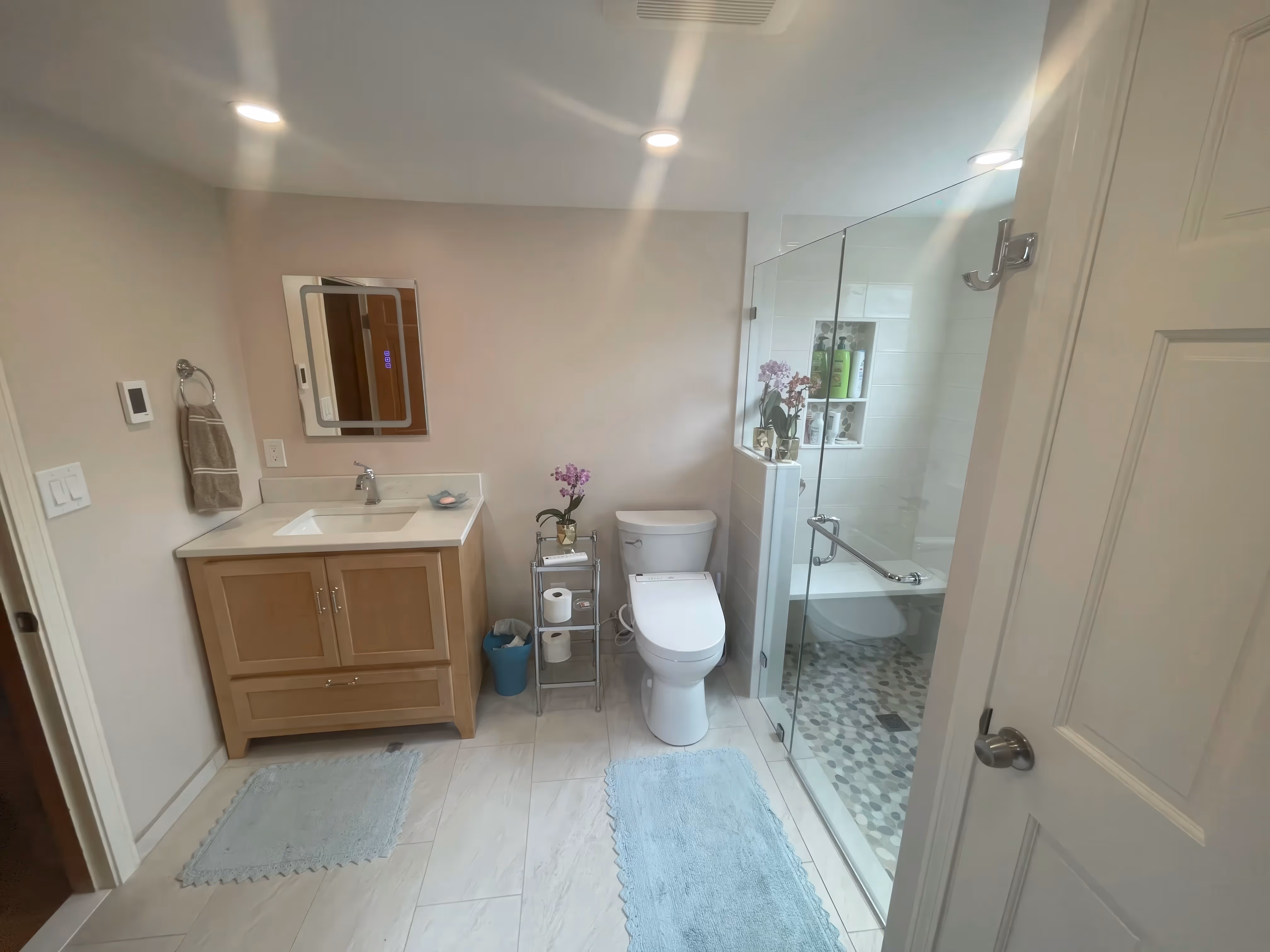 Modern bathroom design with glass walk-in shower, toilet, sink, vanity, orchids, and light blue rugs on a tiled floor.