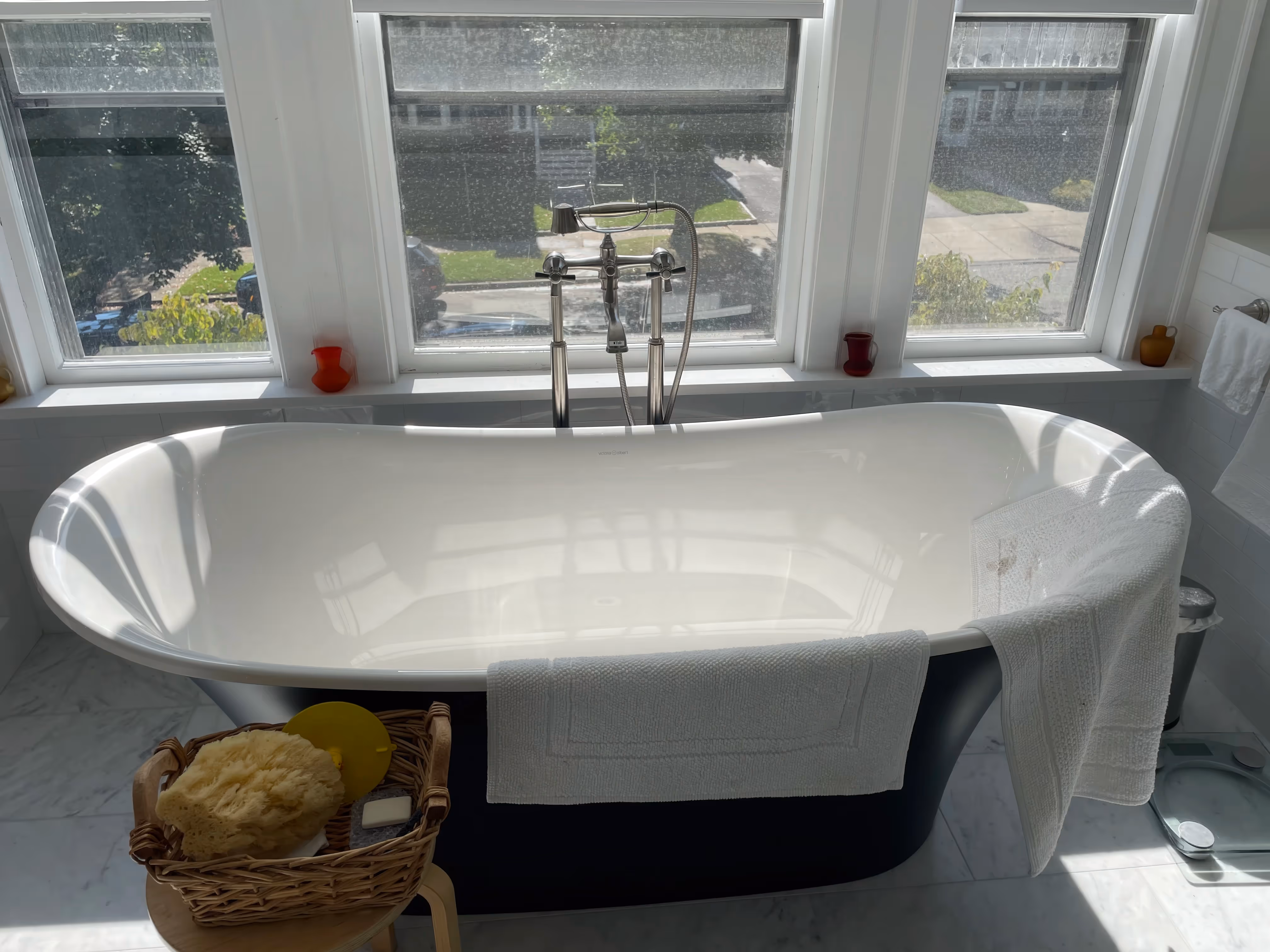 A sunlit bathtub by large windows, with a towel draped over the side, bath items in a basket nearby, and heated floors that add comfort to this inviting bathroom design.
