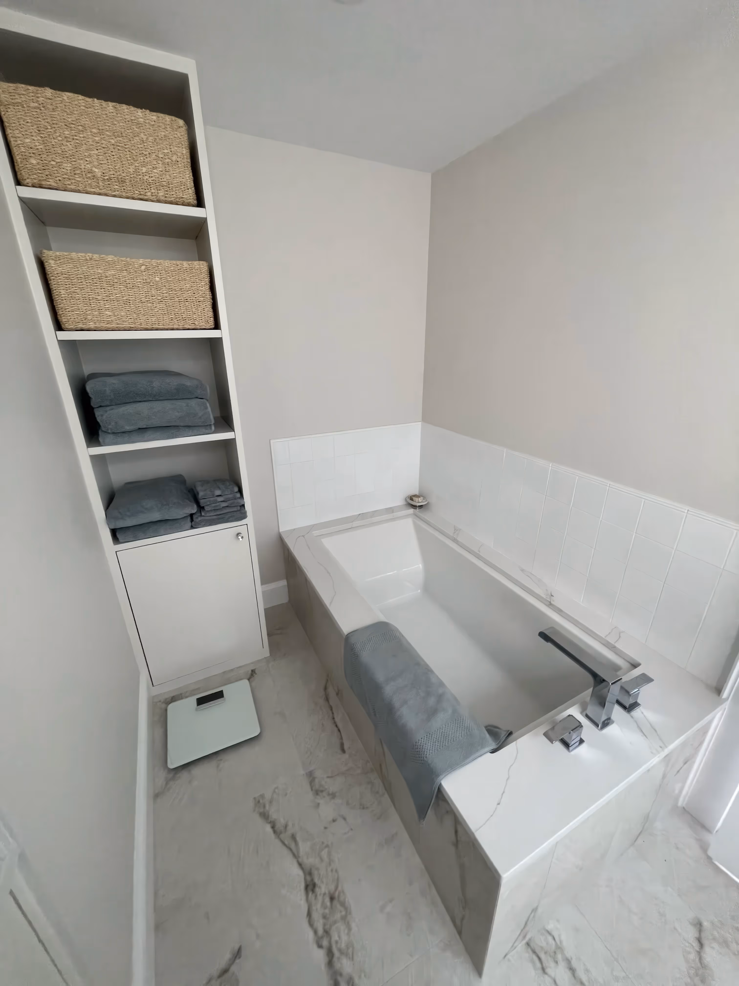 Modern, luxurious bathroom with a white tub, grey towels, storage shelves with baskets, heated floors, and a digital scale on the floor.