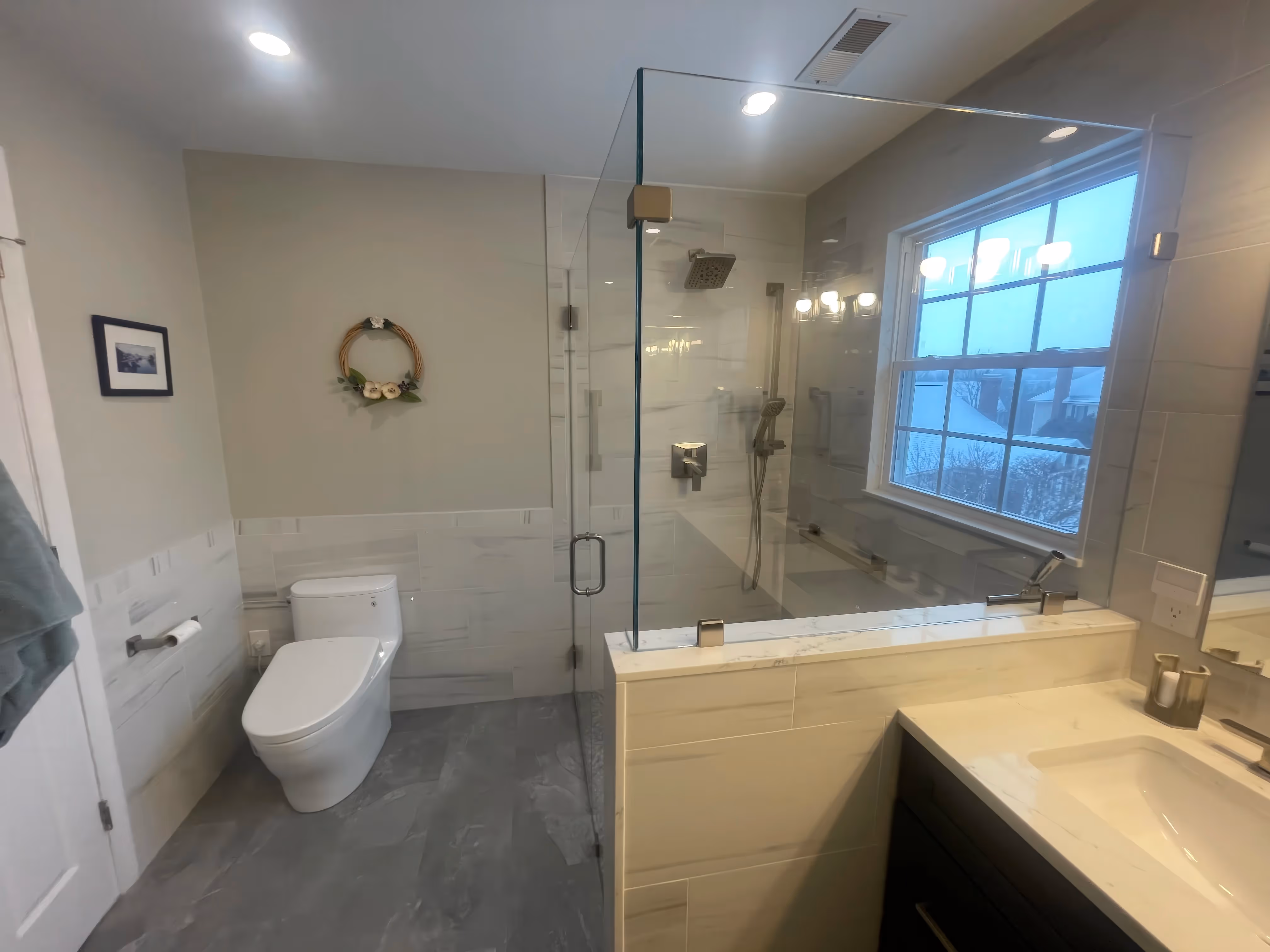 Modern bathroom design with a glass walk-in shower, white toilet, large mirror, and window letting in natural light.