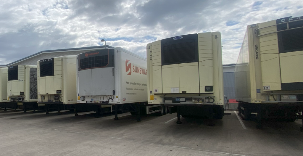 A Sunswap solar and battery-powered transport refrigeration unit (TRU), attached to its trailer, is parked with 4 diesel-powered TRUs and their trailers, 2 either side.