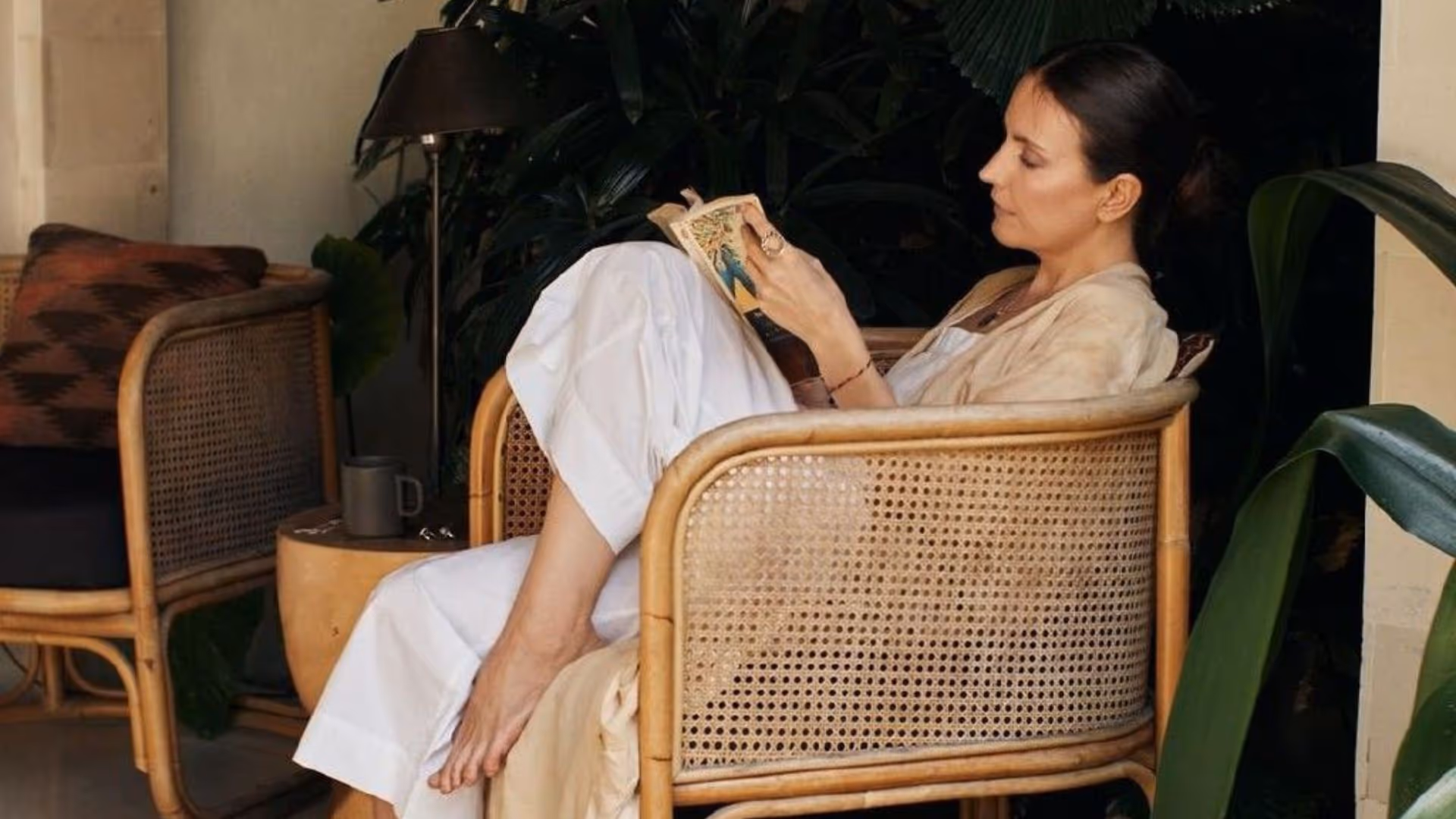 A woman enjoys her leisure time in a comfortable rattan chair, immersed in a book amidst a calming, tropical ambiance.