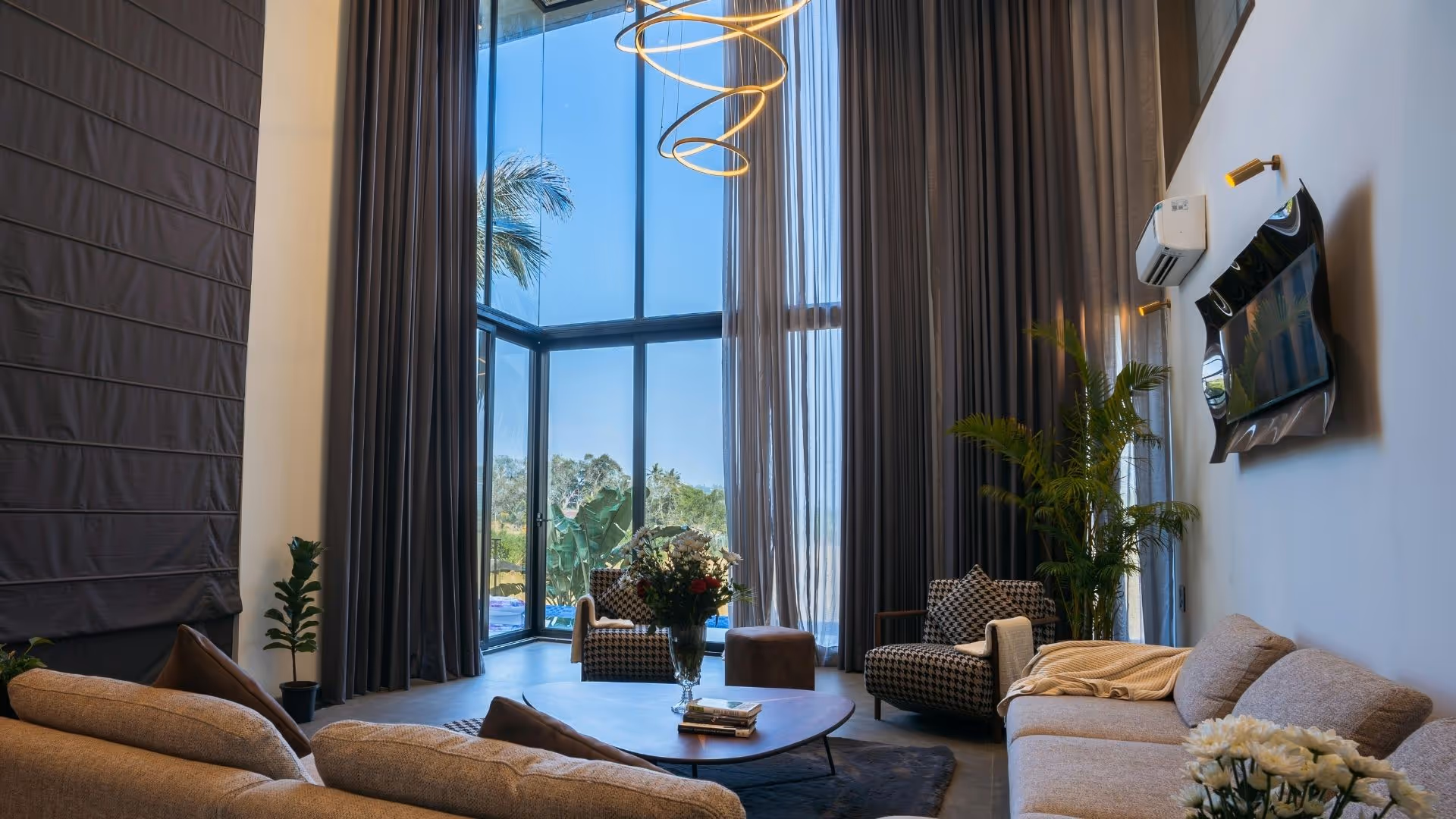 A modern, sunlit living room with floor-to-ceiling windows, a stylish spiral chandelier, and a view of palm trees.