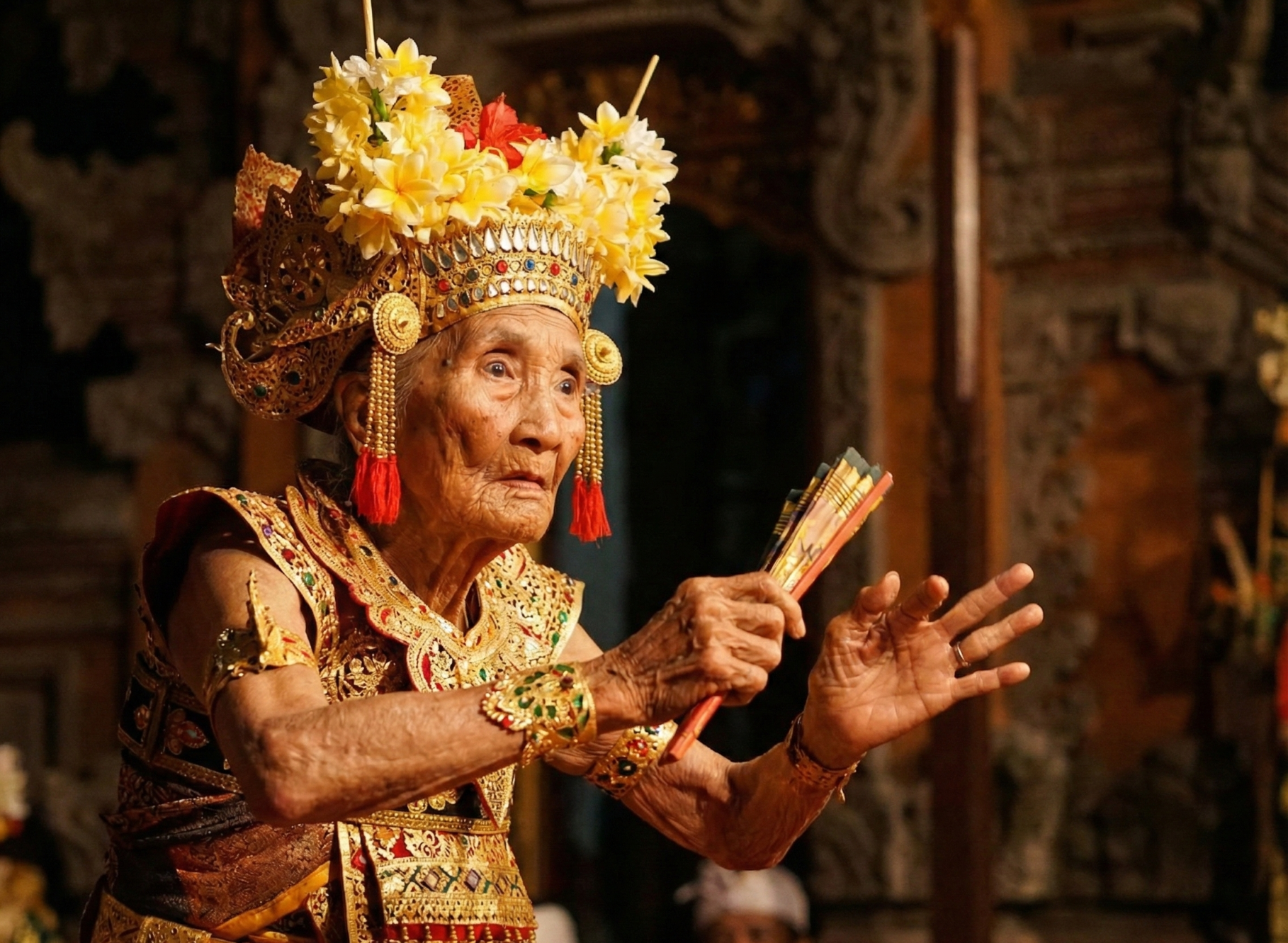 A professional close-up photograph of Niang Soli, the centenarian keeper of the Legong Bapang Saba dance. She is captured mid-performance, wearing an ornate gold traditional costume and an elaborate headdress with fresh flowers, her aged hands holdi