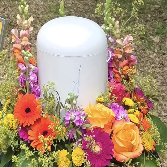 Weiße Urne, umgeben von farbenfrohen Blumen, darunter orangefarbene Gerbera und gelbe Rosen, in Freising.