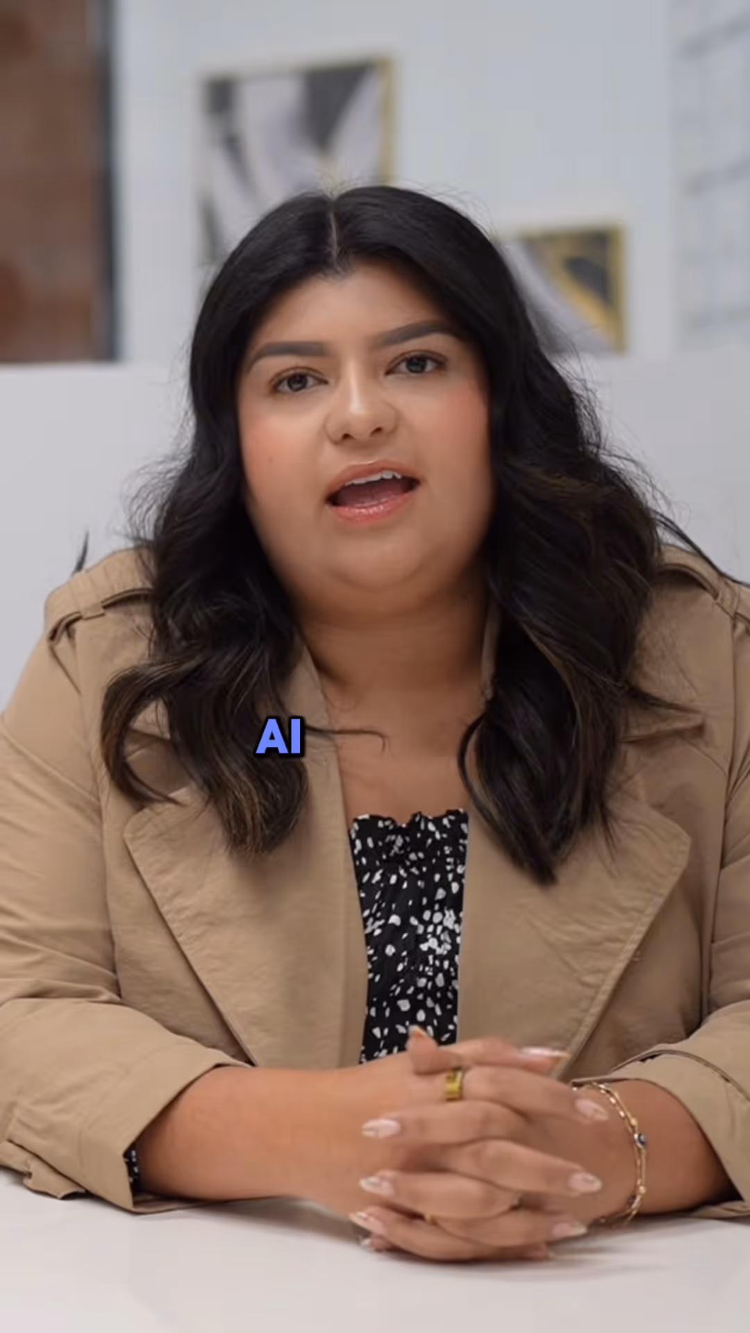 Woman with dark hair and beige jacket speaking with hands clasped on white table.
