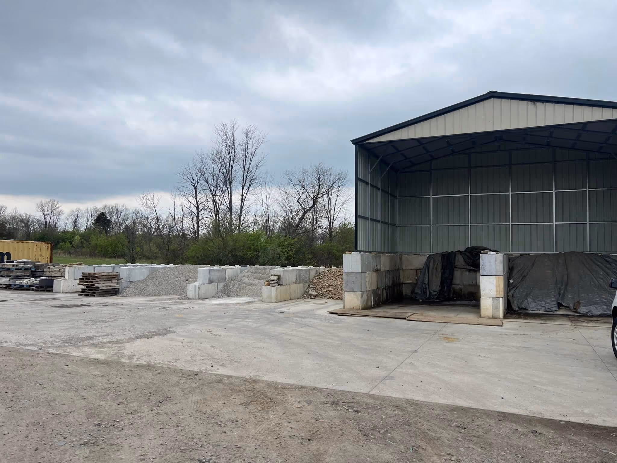 Outdoor storage area with piles of gravel, stone, and other materials separated by concrete block walls, under cloudy sky.