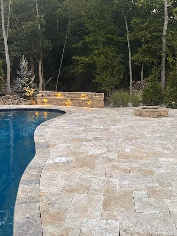 Curved poolside paved with beige tiles, a stone fire pit, and a stone wall with built-in yellow lights surrounded by trees.