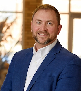 Smiling man with short hair and beard wearing a blue suit jacket and white shirt indoors.